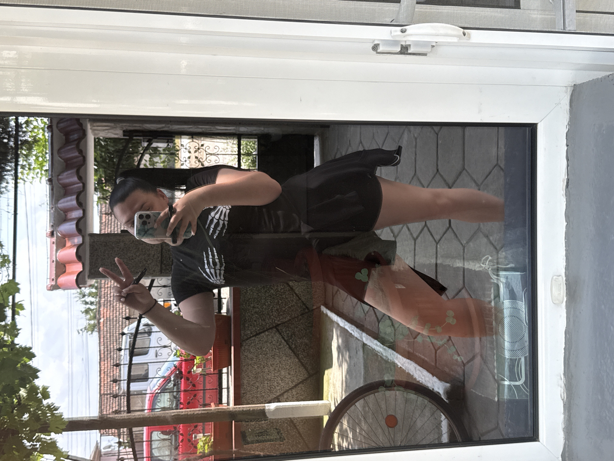 The image captures a person taking a selfie reflected in a large glass window or door. The original image is rotated 90 degrees counter-clockwise.
**Subjects:** The central subject is a person, likely female, with dark hair tied back. They are wearing a black t-shirt with a white skeleton hand design on the front and a black skirt or shorts. The person is holding a smartphone with a patterned case (green, blue, white tones) in their right hand, using it to take the selfie. Their left hand is raised, forming a peace sign (V-sign).
**Scene and Setting:** The person is reflected in what appears to be a clean glass panel set within a white PVC frame, likely part of a window or balcony door. Through and within the reflection, an outdoor scene is visible. This includes parts of a building with red or terracotta roof tiles, lush green foliage and trees, a paved ground surface, and an ornate metal fence. A bicycle wheel is partially visible at the bottom of the reflection. The white PVC frame has a visible white handle/latch mechanism at the top.
**Activity:** The person is actively posing for a selfie, indicated by holding the phone towards the reflection and making a peace sign.
**Notable Details:**
* **Time of day:** The outdoor scene is brightly lit, suggesting it is daytime and sunny, with visible shadows on the ground.
* **Weather:** Clear and bright.
* **Location cues:** The architecture, particularly the style of roof tiles and the general outdoor setting, could be consistent with the given location context of Rogosh, Bulgaria.
* **Emotions:** The peace sign gesture implies a casual, friendly, or relaxed mood.
* **Visible Text:** No legible text is visible on the phone screen or the exterior background. The person's shirt clearly features a skeleton hand design. There's a subtle brand name or model indication on the white window latch, but it's not legible.