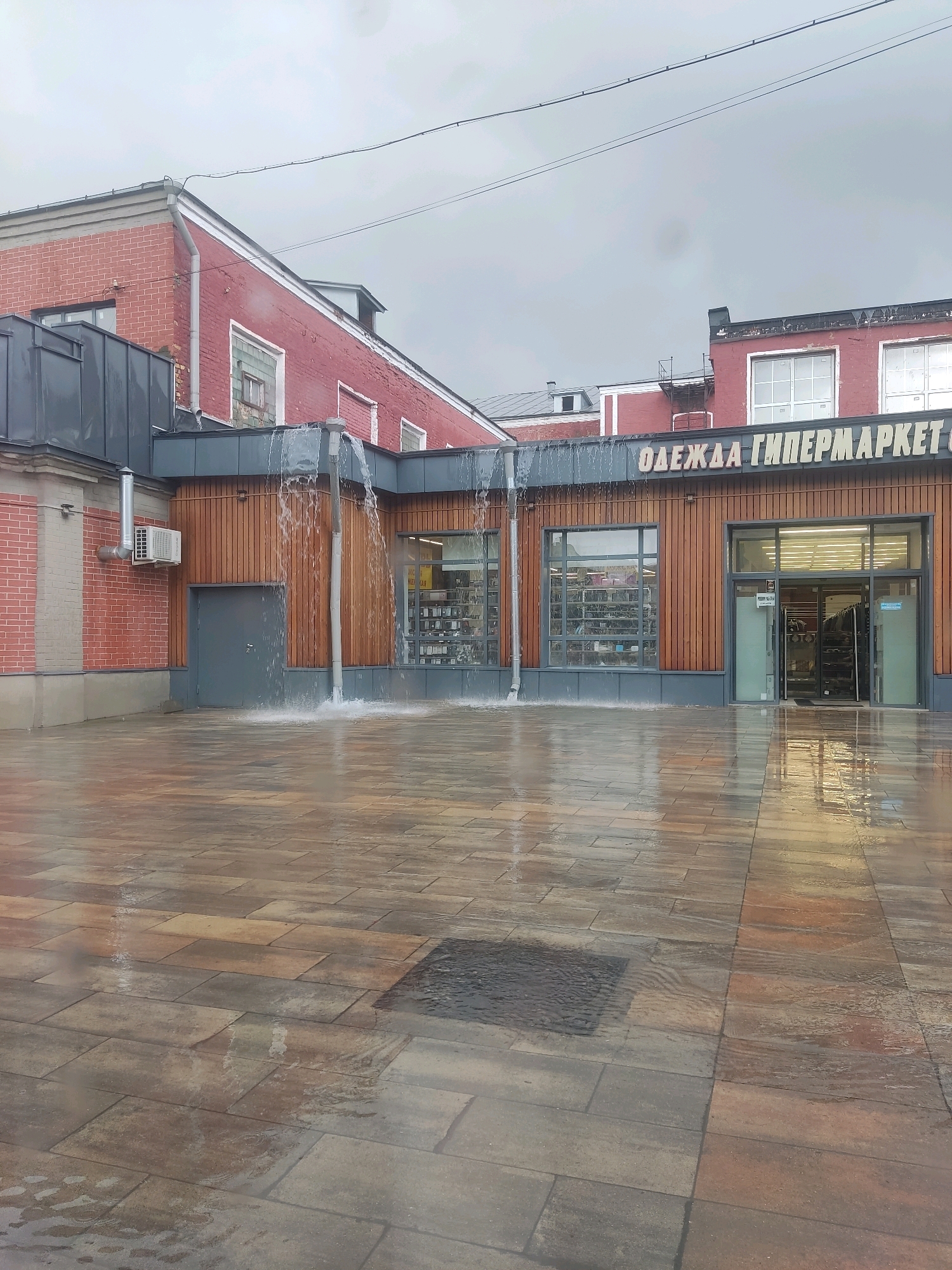 The image depicts the exterior of a shopping center in Noginsk, Russia, during a downpour. Water is gushing from the gutters of the red brick building, creating a waterfall effect onto the paved courtyard. The building features large windows displaying merchandise and a prominent sign that reads "ОДЕЖДА ГИПЕРМАРКЕТ" (Clothing Hypermarket). An air conditioning unit is mounted on the brick wall. The courtyard is paved with large, rectangular stones that are wet and reflective due to the rain. The sky is overcast, indicating a gloomy weather day. No people are visible in the image.