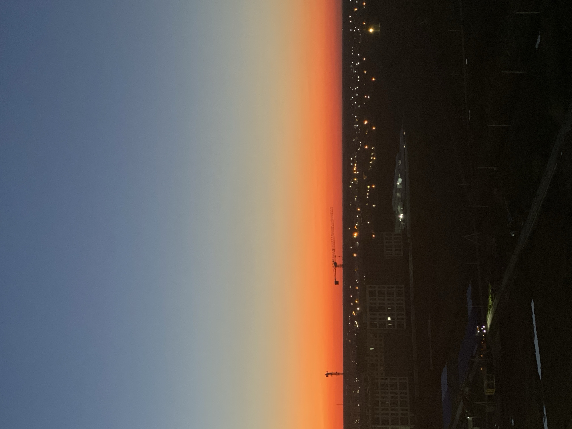 This is an aerial view of a city at dusk, likely Krasnodar, Russia. The sky transitions from a deep blue at the top to a vibrant orange and then a soft yellow near the horizon. Silhouetted against this colorful sky are two construction cranes, one with a red tower and the other a larger yellow lattice crane. In the mid-ground, a cluster of buildings, appearing to be apartment complexes or residential blocks, are visible with lights on in some windows. Further into the distance, the city lights twinkle across the landscape, suggesting a populated urban area. The foreground shows a dark, undeveloped area with some structures and possibly a road. The overall impression is a developing urban landscape at sunset.