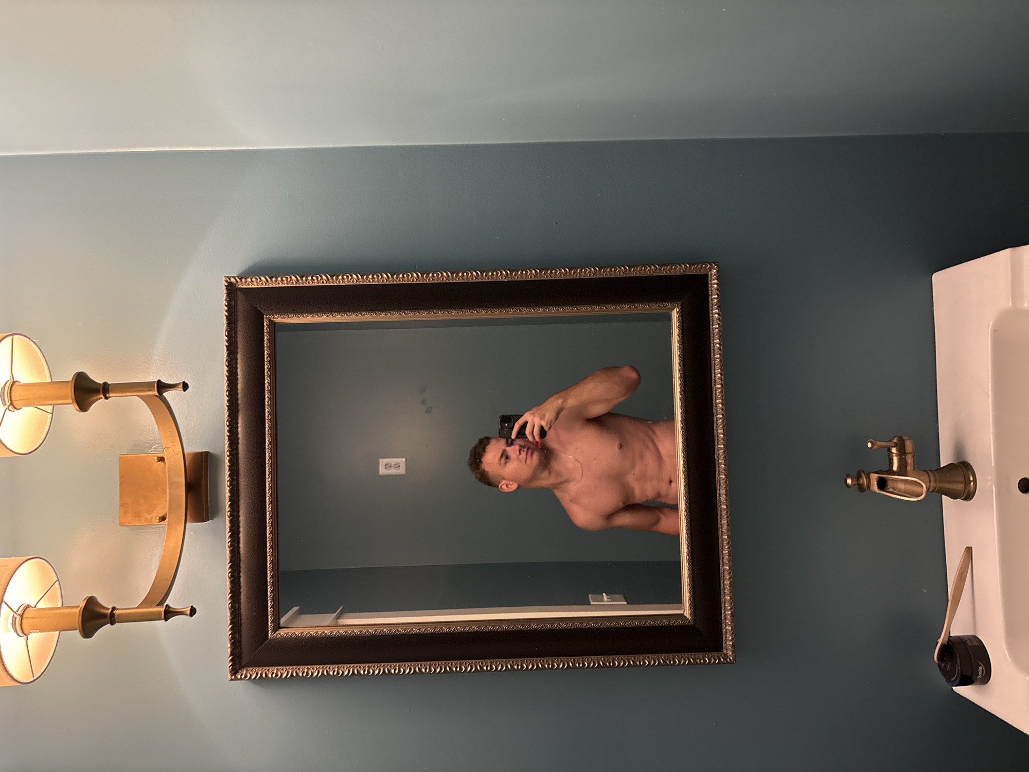 A shirtless person is taking a mirror selfie in a bathroom. The person is standing in front of a large, ornate mirror that is hung on a teal-colored wall. The person is looking at the phone they are holding, which is also visible in the mirror. The person has short, curly hair and is wearing a necklace. In the reflection of the mirror, a light fixture with two shades is visible above the mirror. Below the mirror, a white sink with a brass faucet is visible, along with a toothbrush and a small container on the edge of the sink. The scene appears to be in a residential bathroom. This image was taken in Panama City Beach, United States of America.