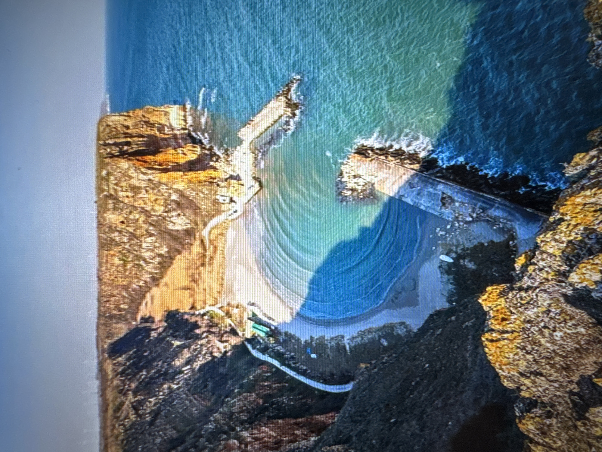 An aerial view captures a rugged coastal cove, likely a secluded beach, framed by steep, rocky cliffs. The image is taken from above, looking down into the bay. The main feature is a crescent-shaped sandy beach, meeting clear, blue-green ocean water. Visible patterns of waves or currents are present in the water, particularly near the shore. A prominent dark shadow extends across part of the water and beach, suggesting an overhead feature like a cloud or a higher part of the cliff out of frame.
The cliffs flanking the cove are a mix of earthy tones, including golden browns and dark grays, indicating rock formations with some sparse vegetation. Man-made structures are visible: two concrete or stone breakwaters extend into the sea, defining the entrance to the cove. A light-colored, winding path or road can be seen descending from the top of the cliff on the left side down to the beach. At the base of the cliffs, near the sand, there are several small, simple structures, possibly shelters or boathouses, some with green or blue roofs. A few white, small objects, likely boats or kayaks, are scattered on the sandy shore.
The scene is brightly illuminated by natural daylight, casting distinct shadows. No people or specific activities are discernible, giving the area a tranquil appearance, despite evidence of human presence and utilization. The image itself appears to be a photograph of a digital screen, showing noticeable pixelation and scan lines.