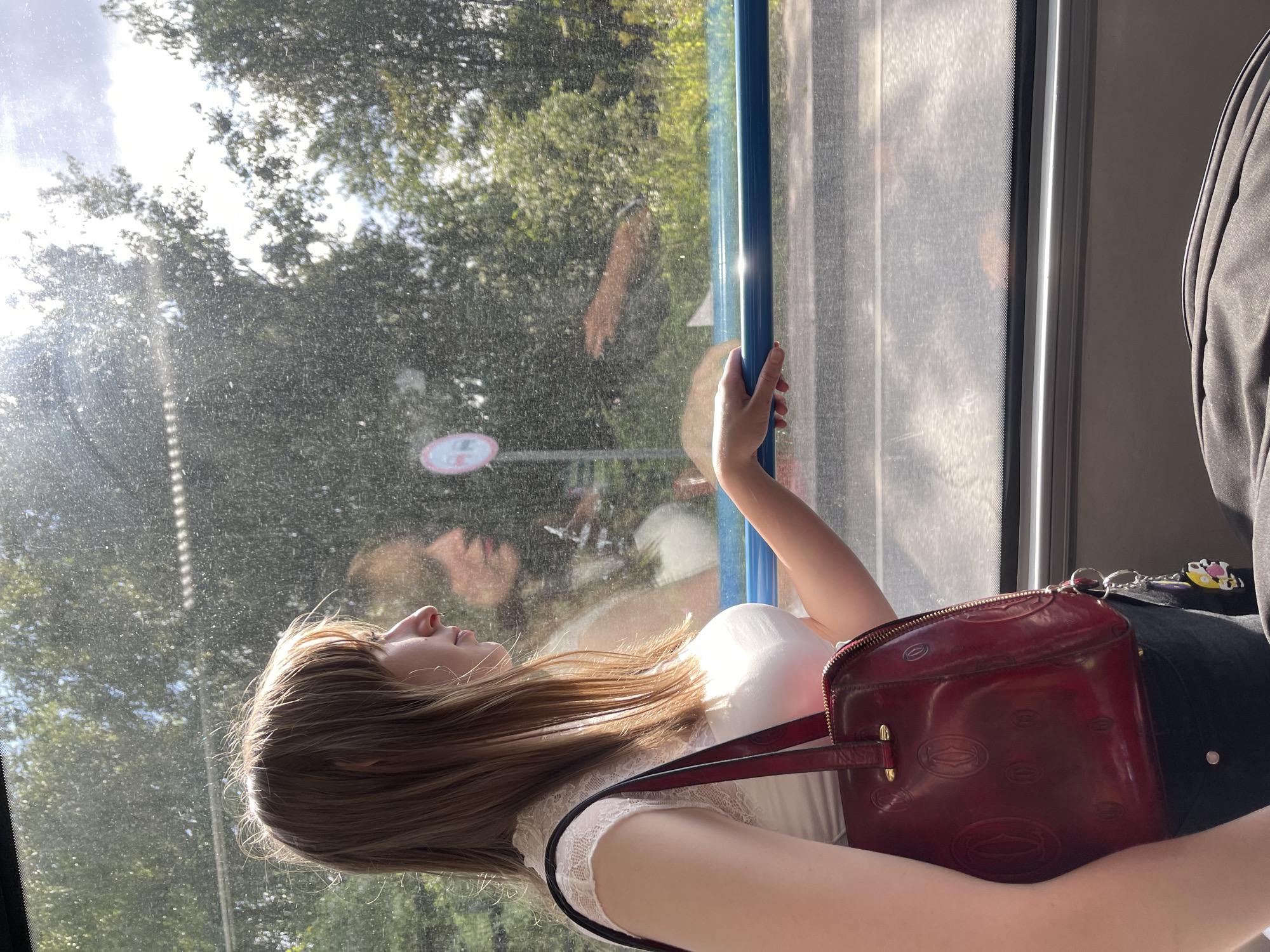 A young woman with light brown hair and bangs is seated by a window, looking out. She is wearing a white lace-trimmed top and black pants, and is carrying a red handbag. Her hand rests on a blue bar along the window. The window is dirty and reflects the interior of what appears to be a bus or train, with a fuzzy image of another person visible. Outside the window, trees and a blue sky with clouds can be seen. A round red and white sign is also visible through the window. The overall lighting suggests a sunny day. The location is identified as Sanatoriya "Energiya", Russia.
