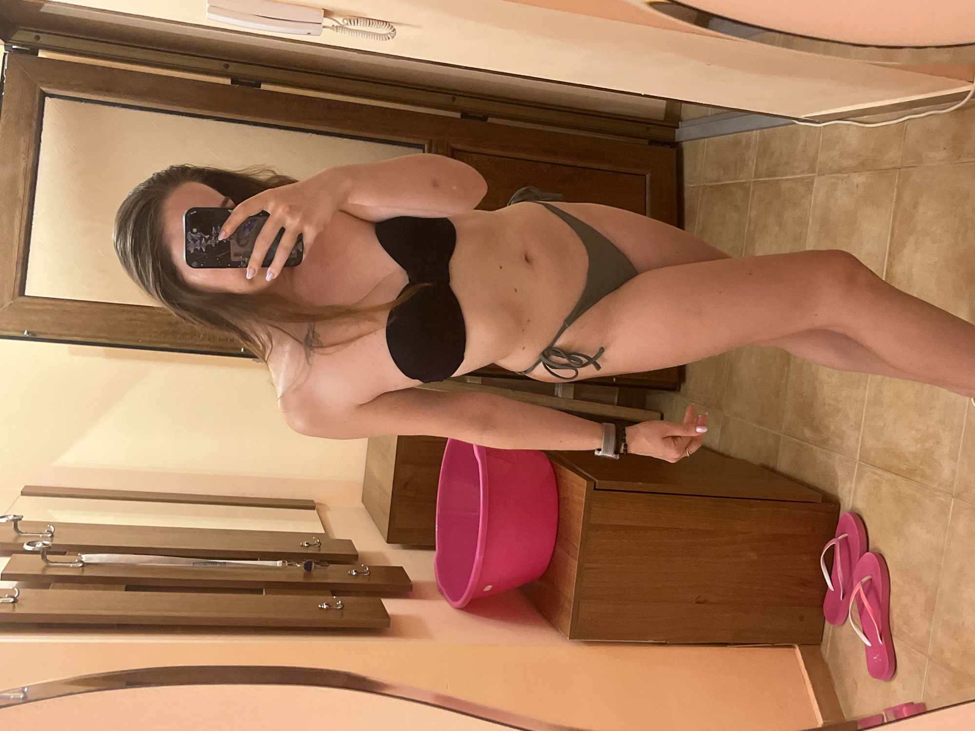 A woman stands in a changing room and takes a selfie in a mirror. She is wearing a black strapless bikini top and olive green bikini bottoms. The room has tiled floors and peach-colored walls. To the left of the woman, there is a wooden cabinet with a pink basin on top. To the right, there is a wooden door and a wall-mounted telephone. There are also hooks on the wall, possibly for hanging clothes. In the foreground, a pair of pink flip-flops are on the floor. The image does not provide enough information to determine the specific location in Lozenets, Bulgaria.