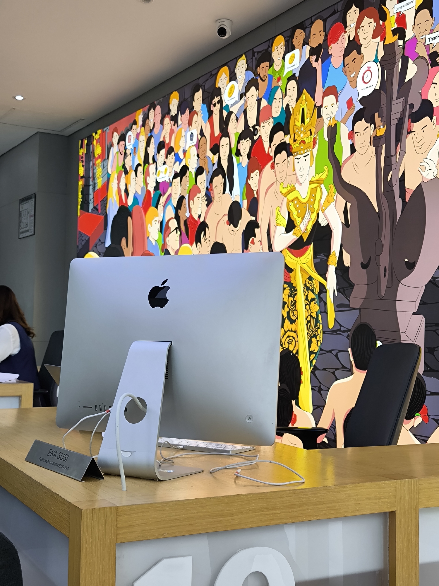 The image depicts an office setting in North Kuta, Indonesia, with a focus on a desk area. The desk is made of light-colored wood and features an iMac computer with an Apple logo prominently visible on the back. A nameplate on the desk identifies "Eka Susi" as a Customer Experience Officer. A white keyboard and a tangle of wires are on the desk, along with a curved metal stand supporting the computer.
Behind the desk, a large, colorful cartoon mural dominates the background. The mural depicts a diverse crowd of people, some holding speech bubbles with various messages. Central to the mural is a figure dressed in traditional Indonesian attire, including an ornate headdress and yellow patterned clothing. Also depicted is part of a dark gray elephant sculpture.
To the left of the desk, a person with dark hair is sitting, only partially visible. A security camera is mounted on the ceiling. Large white numbers are on the front of the desk.