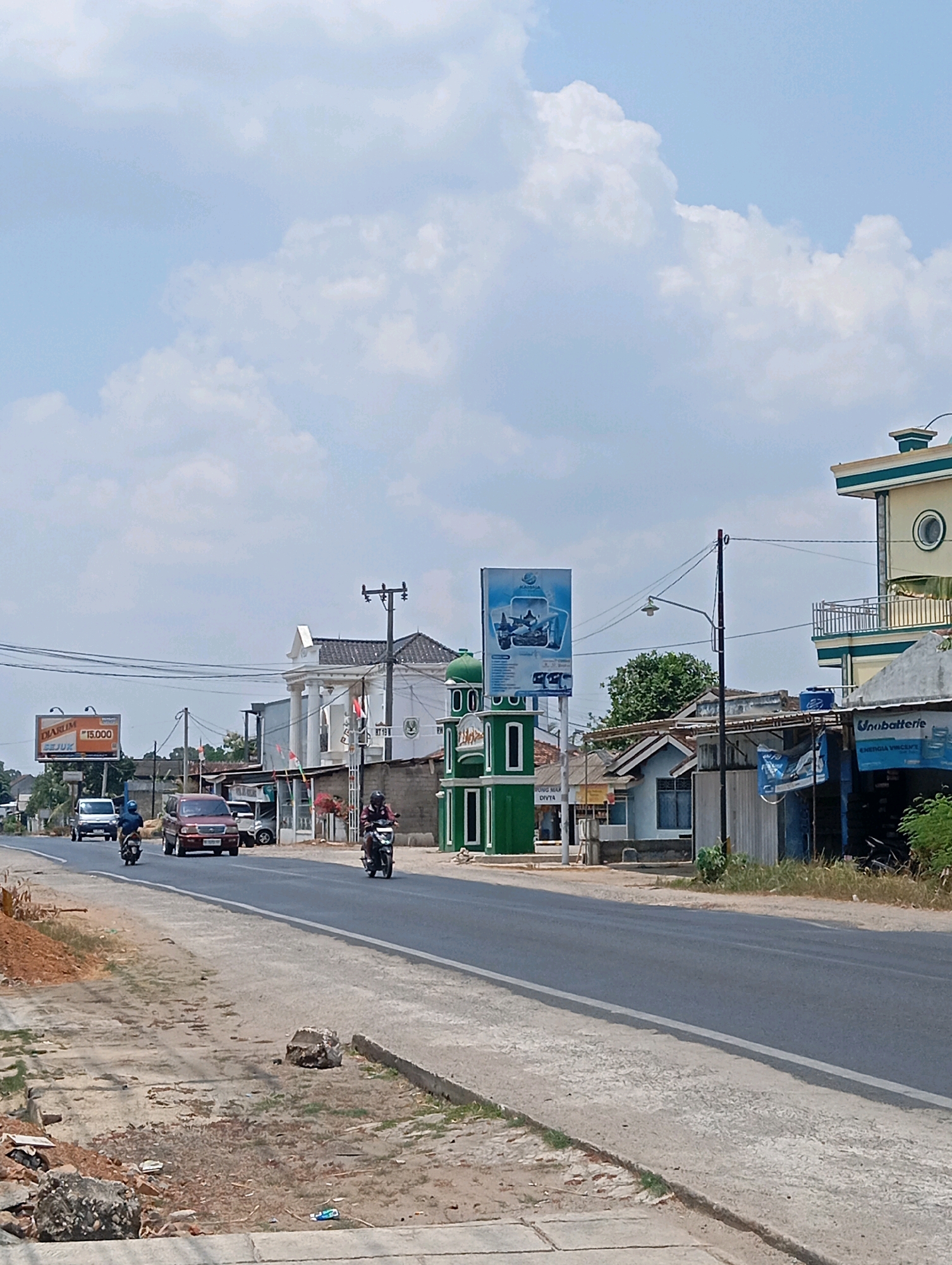 In the city of Leman Benawi, Indonesia, a two-lane asphalt road runs through a semi-urban area. On the left side of the road, there's a dirt shoulder with piles of dirt and debris, leading to a large billboard advertising "Djarum" cigarettes. Several vehicles, including a white van, a maroon SUV, and two motorcycles with riders, are traveling on the road.
The right side of the road features a mix of buildings and vegetation. A prominent green structure resembling a minaret stands near the roadside. Further down, there's a two-story building with a balcony and a distinctive circular window. Adjacent to it are several single-story commercial establishments, one with a sign that reads "Unabatterie." Power lines and a streetlamp are visible overhead, indicating the presence of infrastructure. The sky is partly cloudy with a bright, hazy atmosphere, suggesting daytime. The scene appears to be a typical commercial street in a developing urban or semi-urban environment.