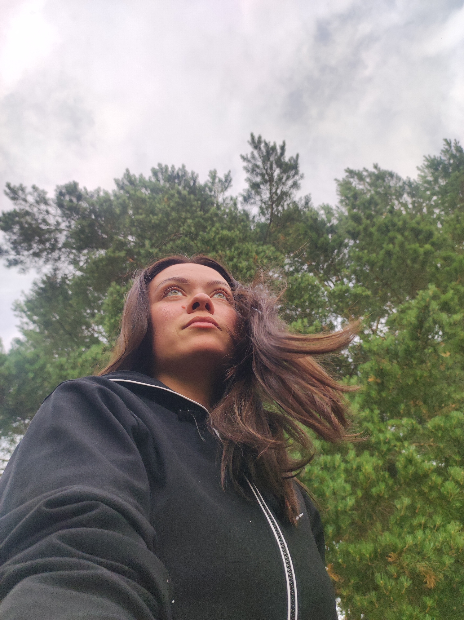 A low-angle, medium shot captures a young woman looking up at the sky, with a blurred background of pine trees and an overcast sky. She is wearing a black zip-up hoodie with white trim around the zipper. Her brown hair is blowing across her face. The image appears to have been taken outdoors in a natural setting.