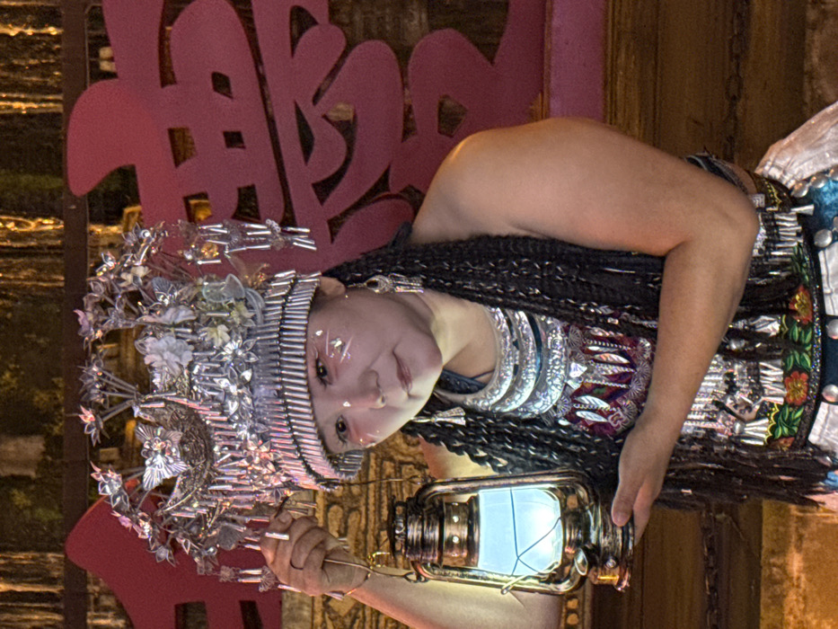 An adult female, likely of East Asian descent, is depicted posing in traditional ceremonial attire in Yuzhong District, China. She wears an elaborate silver-colored metal headdress adorned with detailed bird figures, floral designs, and numerous dangling elements. Multiple layers of thick, ornate silver-colored necklaces and matching long earrings are visible. Her face features light makeup, including white lines and dots on her cheeks. Her dark hair is styled in long braids. She holds a bronze-colored hurricane lantern, emitting a distinct light blue glow, with both hands at chest level. Her clothing consists of a dark sleeveless top beneath an elaborately embroidered garment featuring colorful patterns of feathers and flowers. The background is dark and out of focus, showcasing a large, red cutout Chinese character, resembling "囍" (double happiness), to the right, with blurred warm lights visible higher in the frame.