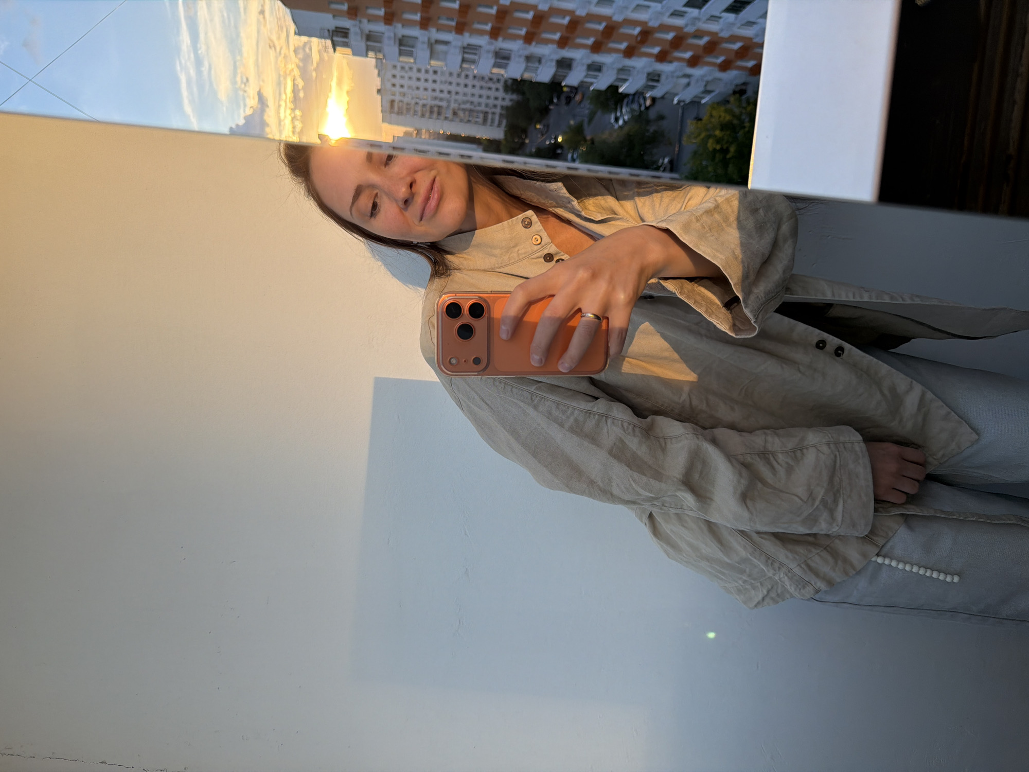 A portrait-oriented photo shows a woman taking a selfie in a reflection, likely from a window or large mirror. The woman, with dark hair, appears to be in an indoor setting, while the reflection reveals an outdoor urban landscape with a sunset.
The primary subject is a young woman, captured from the chest up, with a soft smile and relaxed expression. She is holding an orange/coral-cased smartphone in her right hand, which has a simple metallic ring on her ring finger. She wears a loose-fitting, light beige or off-white button-up shirt or jacket. Her left hand is visible further down, resting near her side, and she appears to be wearing matching or coordinating light-colored pants featuring a decorative string of white beads along a seam or pocket.
The reflection behind her head and shoulders prominently displays a golden sunset over an urban environment. Tall apartment buildings with multiple windows and balconies are visible, characteristic of city architecture in places like Khimki, Russia. Trees and what appear to be parked cars are also discernible in the reflection below the buildings. The sky is partially cloudy, illuminated by the warm, intense light of the setting sun.
The immediate foreground and left side of the image show a plain, light-colored wall, possibly off-white or beige, with some minor scuffs and faint cracks at the very bottom. The overall lighting suggests late afternoon or early evening due to the sunset. No visible text is present in the image. The mood is calm and serene.