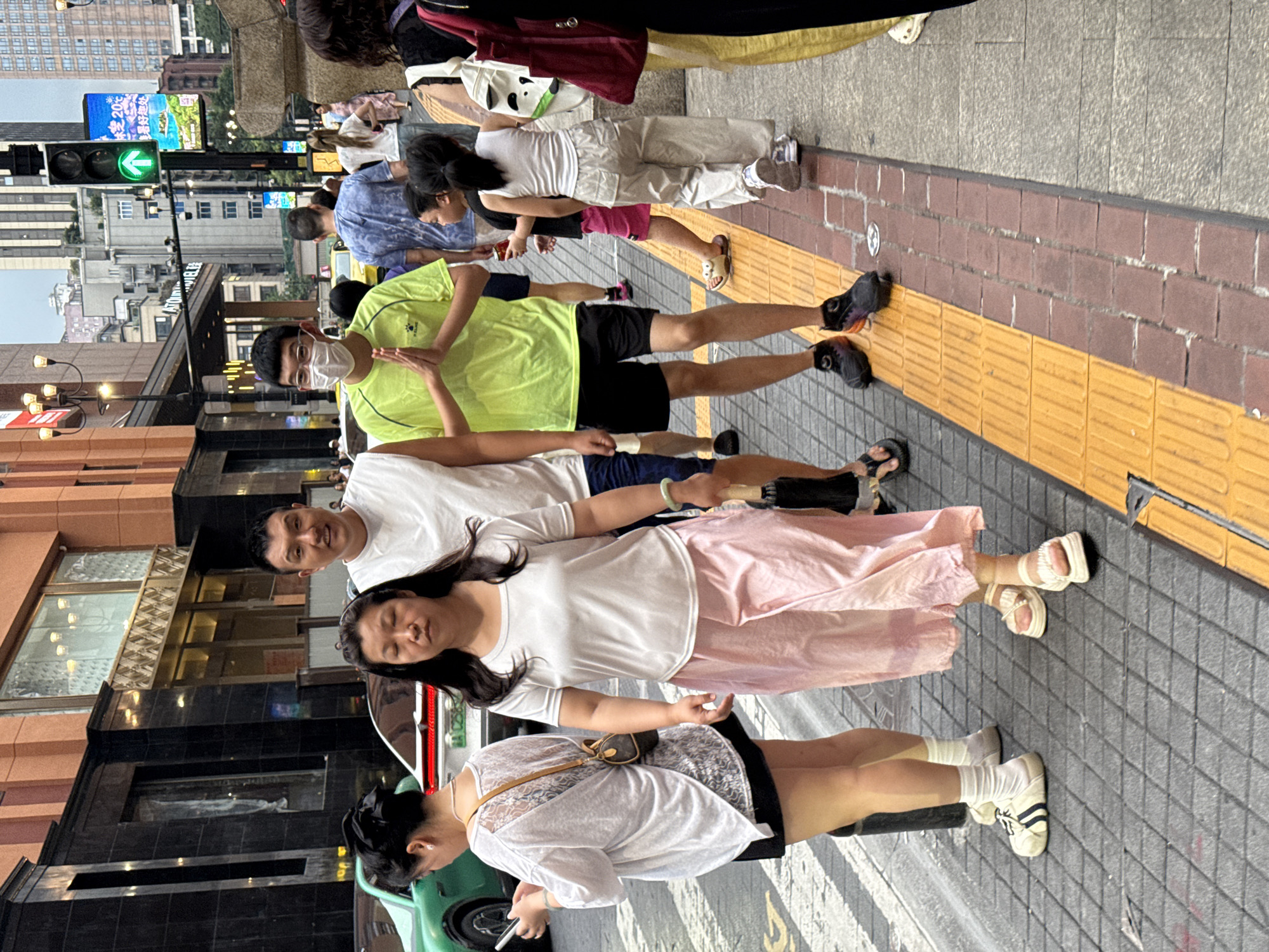 In Yuzhong District, China, a group of people are walking on a city sidewalk next to a street. In the foreground, a woman with dark hair walks towards the viewer, wearing a white t-shirt and a pink skirt. To her left, another woman, facing away, wears a white sheer top over a black skirt and carries a small brown purse. A man in a white t-shirt and dark shorts stands behind the woman in the pink skirt, looking towards the viewer. To his right, a man wearing a neon green t-shirt and black shorts stands with his hands pressed together in front of him. Further back, a young girl in a white tank top and beige pants walks away from the viewer, and other pedestrians and vehicles are visible on the street and sidewalk. The environment includes buildings with modern architecture, a traffic light displaying a green arrow, and street signage. The ground is paved with gray tiles and features a tactile paving strip.