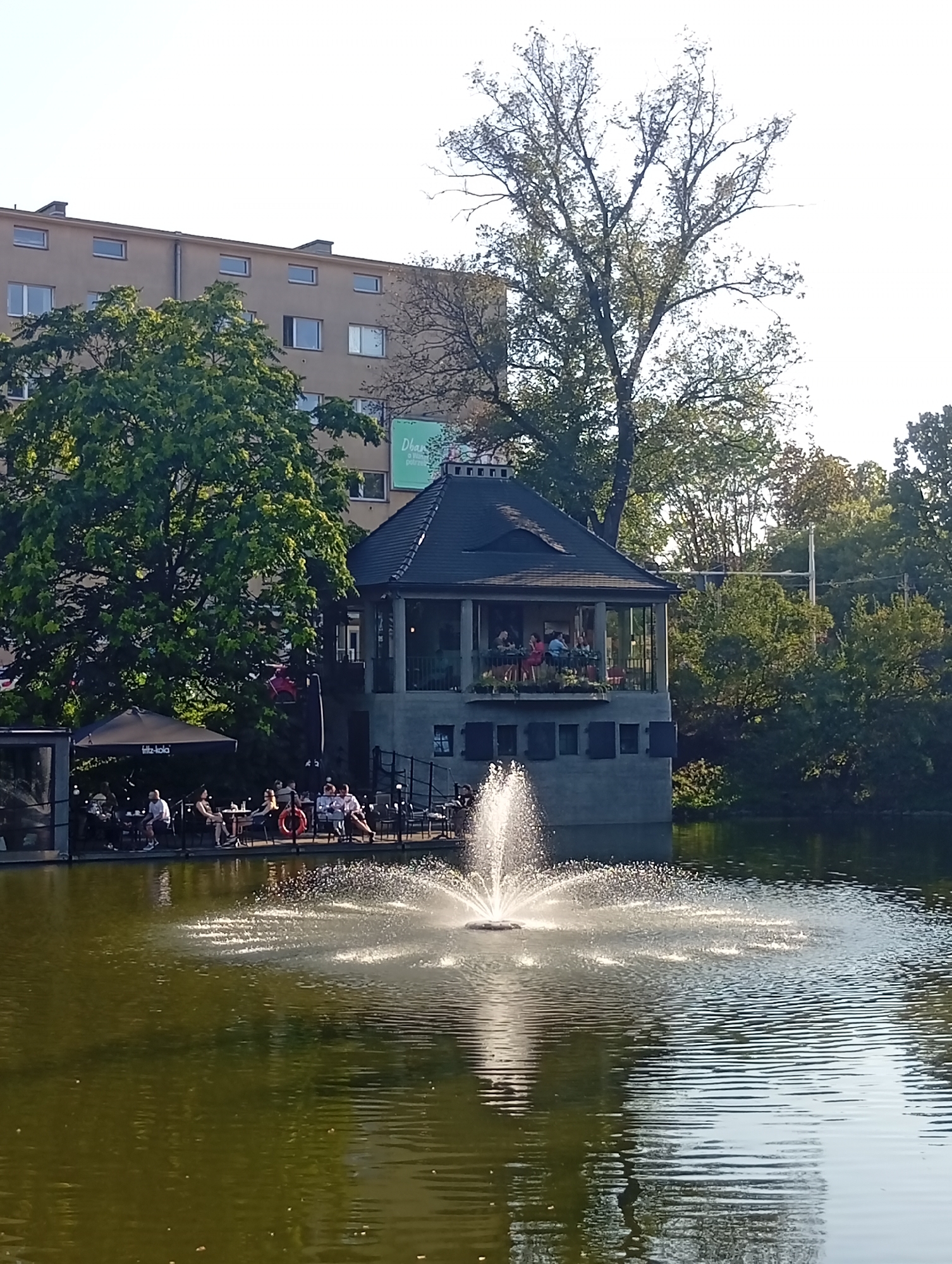 The image depicts a sunlit outdoor cafe situated on the edge of a body of water, likely a pond or lake, in Wrocław, Poland. A prominent fountain sprays water in the foreground, creating a shimmering effect on the water's surface. In the mid-ground, a multi-story building with a dark, conical roof and a glass-enclosed upper level overlooks the water. Several people are seated at tables on a patio area adjacent to the building, some under a black umbrella with "fritz-kola" branding. The atmosphere appears relaxed, with individuals engaged in conversation and enjoying the outdoor setting. In the background, a large, mature tree with lush green foliage stands to the left, and further behind it, a modern, beige apartment building with numerous windows is visible. The overall scene captures a pleasant day at a waterside establishment in an urban environment.