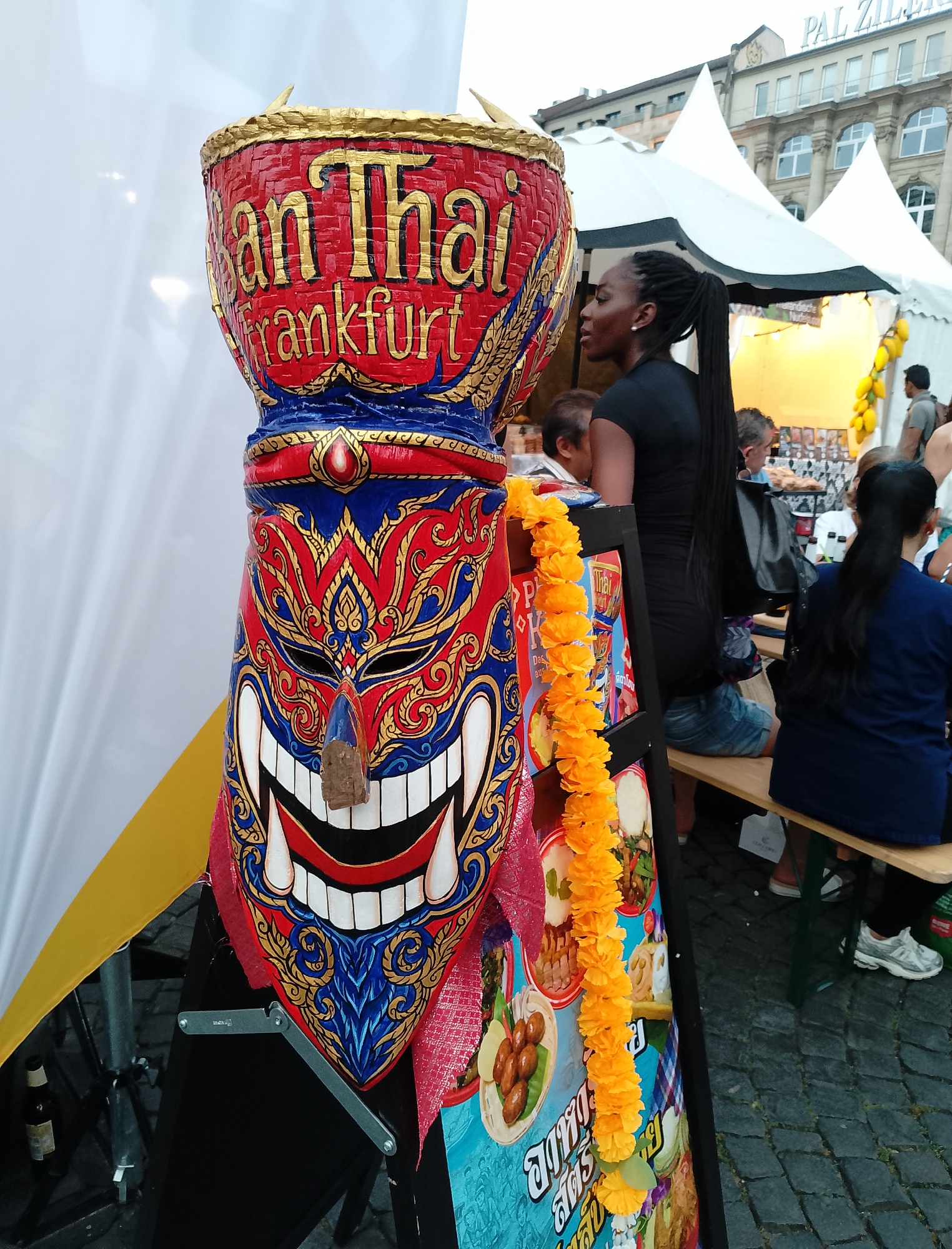 A large, brightly colored, mask-like wooden sculpture stands in the foreground, adorned with gold and blue intricate patterns and a wide, grinning mouth. A yellow garland of flowers hangs to its right. Behind the sculpture, several people are visible, including a Black woman with braided hair wearing a black dress, and other individuals seated at wooden tables outside of white tents. The background shows buildings with arched windows and a sign reading "PAL ZI..." atop one of them. The ground is paved with cobblestones. The scene appears to be an outdoor market or festival.