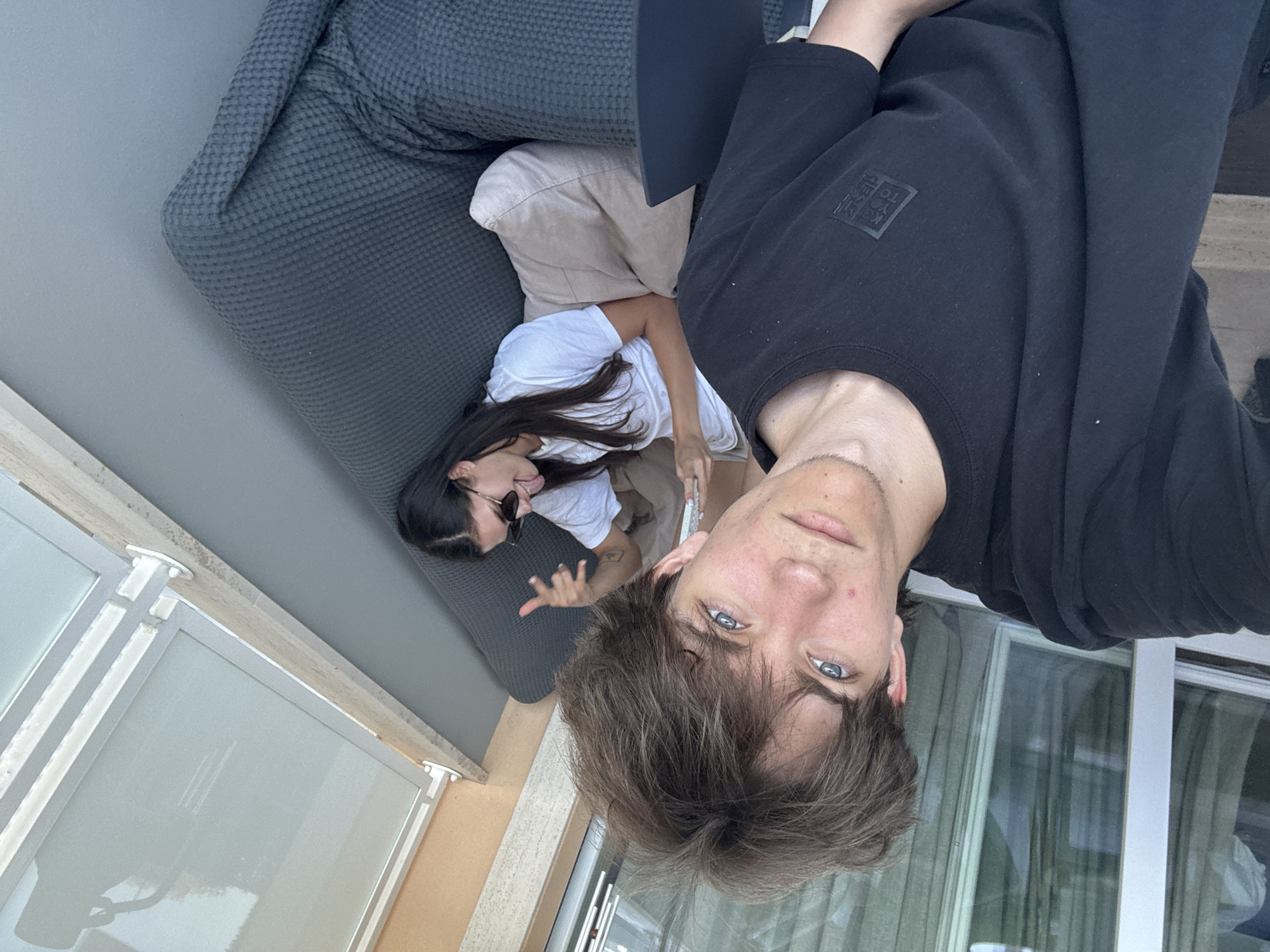 A young man with brown hair and blue eyes is taking a selfie on a balcony, wearing a black t-shirt. Beside him, a young woman with dark hair is lying on a dark grey couch, wearing a white t-shirt and beige pants. She is sticking her tongue out and making a peace sign with her fingers. The balcony has a light-colored railing and a large window with vertical blinds. In the background, there are views of buildings and greenery, suggesting an outdoor urban or semi-urban setting, possibly a residential area in Cupra Marittima, Italy, given the architecture and landscaping. The overall mood of the image is casual and playful.