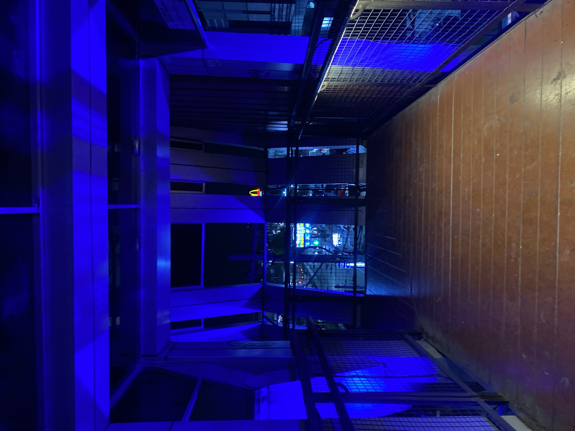 The image depicts a pedestrian bridge at night, illuminated by vibrant blue lighting. The bridge has a wooden walkway with metal railings on either side. Beyond the railings, the city of Banjarbaru, Indonesia, is visible with illuminated buildings and streetlights. A McDonald's sign with its iconic golden arches is visible in the distance. The scene appears to be a public space designed for pedestrian traffic, with architectural elements that suggest a modern urban setting. There are no people or animals visible in the image.