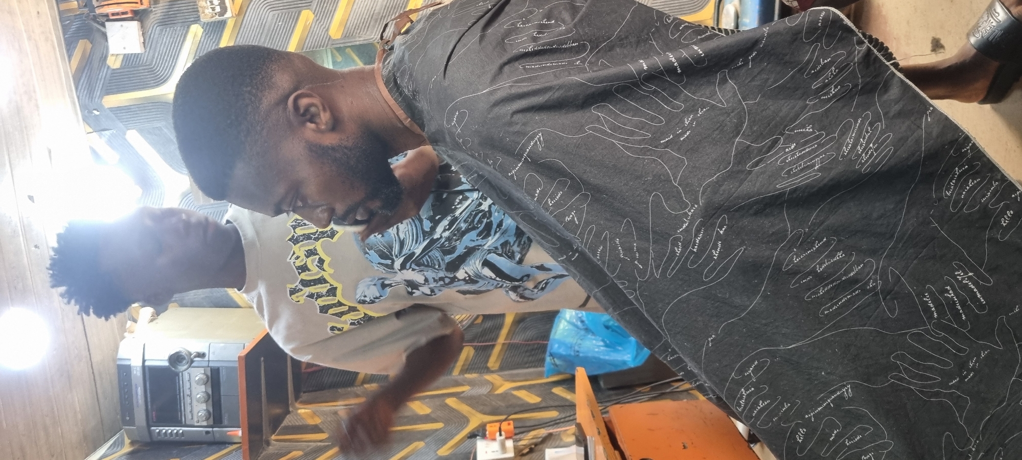 In a barbershop located in Ikeja, Nigeria, a young Black man with short, styled hair and a light-colored t-shirt with a graphic design stands to the left, partially obscured by another individual. He appears to be holding a tool, possibly a clipper, and looking downwards. To the right, a Black man with a beard and a faded haircut sits, wearing a dark barber cape with a white line-art pattern of hands and text. The cape is draped over his shoulders and lap. The background shows a wall with a yellow and grey striped pattern, a shelf with a stereo system, and a bright light source overhead. A blue plastic bag is visible on the left side of the image.