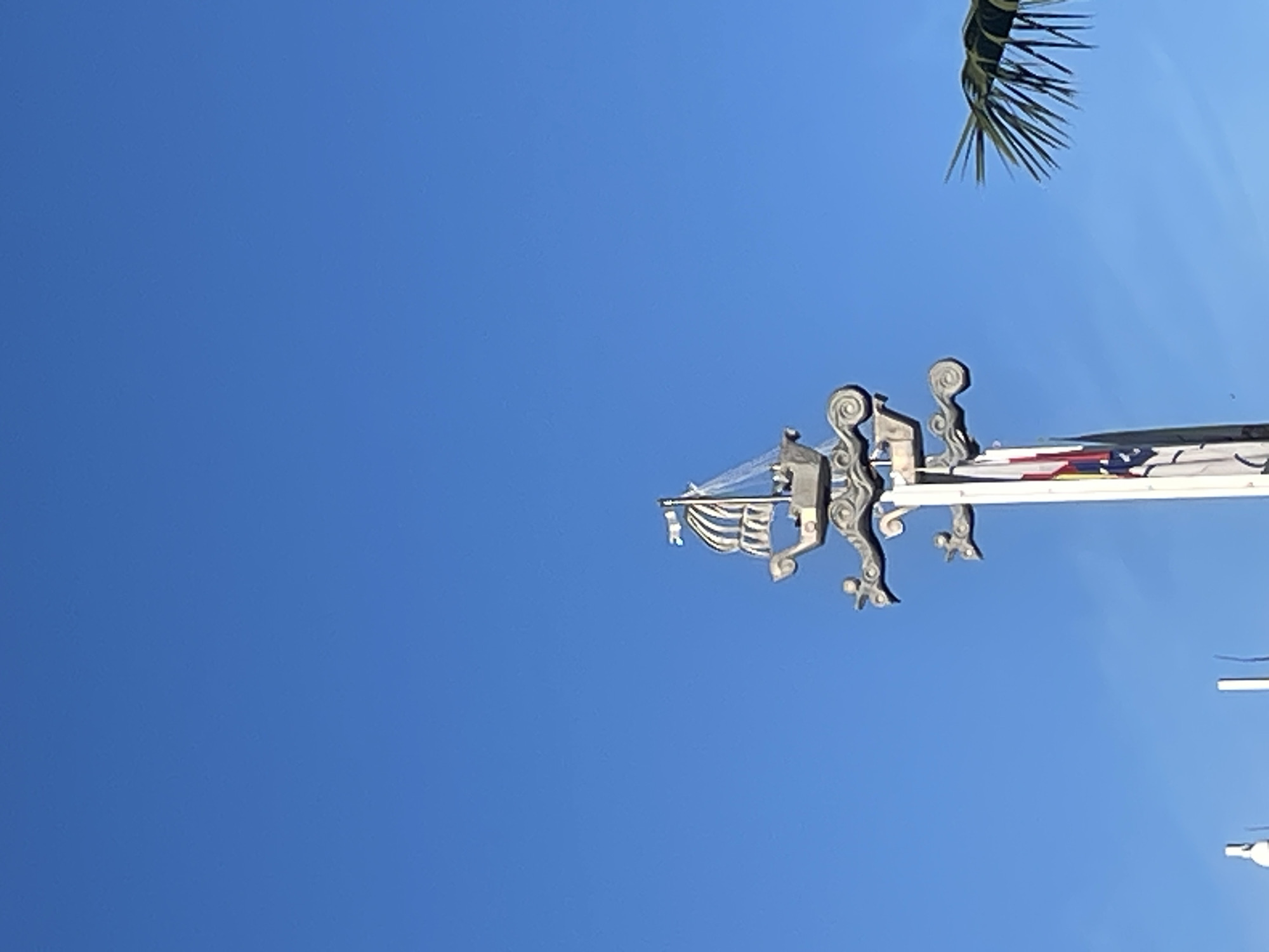 The image displays an ornate architectural detail set against a vibrant, clear blue sky. The main subject is a decorative structure, possibly made of light-colored stone or metal, characterized by intricate scrollwork and possibly stylized figures, oriented vertically along the right side of the frame. Attached to its left side is a smaller, stylized element resembling a sailing ship, complete with a mast and decorative 'sails' or banners. Further along the main structure to the right, a horizontal bar supports what appear to be colorful flags or banners, displaying segments of red, yellow, and blue.
In the upper right corner of the frame, a segment of a green palm frond is visible, suggesting a warm, possibly subtropical, climate. A small portion of a white pole and a spherical object can be seen in the bottom right corner.
The scene is captured during bright daylight hours, indicated by the clear sky and crisp lighting, signifying sunny weather. There are no people visible, and no specific activity or event is depicted, focusing instead on the architectural and natural elements. No visible text is present. The architectural style combined with the presence of palm trees is consistent with the provided location context of Sochi, Russia.