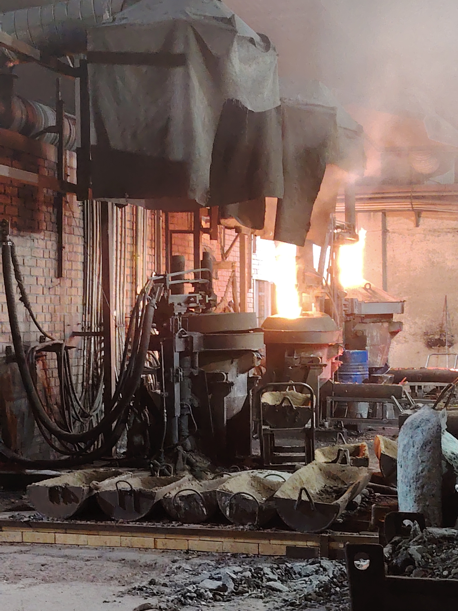 The image depicts the interior of a foundry in Noviy, Russia. Molten metal emits bright light and smoke from two industrial crucibles. Heavy, dark-colored machinery is positioned around the crucibles. Metal buckets, filled with debris, sit in the foreground. A brick wall is visible in the background on the left. Overhead, canvas-covered structures are suspended. The floor is cluttered with loose materials.