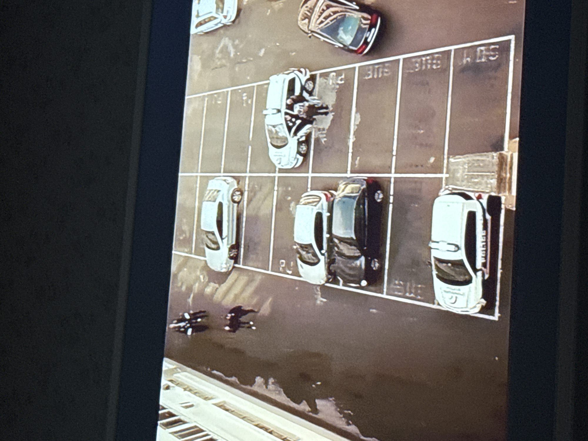 An aerial view of a parking lot in Mytishchi, Russia, features parked vehicles, parking space identifiers, and several figures, likely people. The environment suggests an urban setting. The parking lot layout is visible, with distinct parking spaces. A few vehicles are parked. Some figures are present in the image, although their specific actions or identities are unclear.