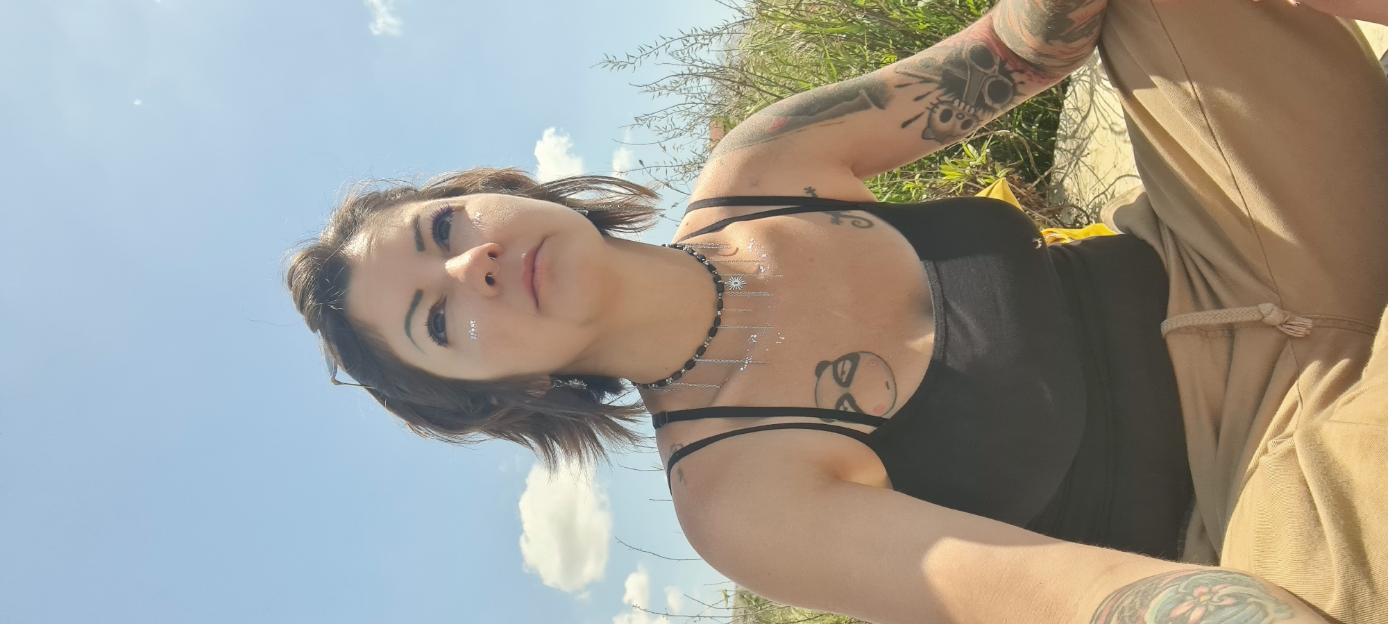 A woman with tattoos is posing outdoors in a sunny location. She is wearing a black tank top and khaki pants. She has short brown hair, a nose ring, and sparkles on her face and chest. A black and silver necklace adorns her neck. The sky is blue with scattered clouds. The background includes green foliage. The city is Lachish Regional Council, Israel.