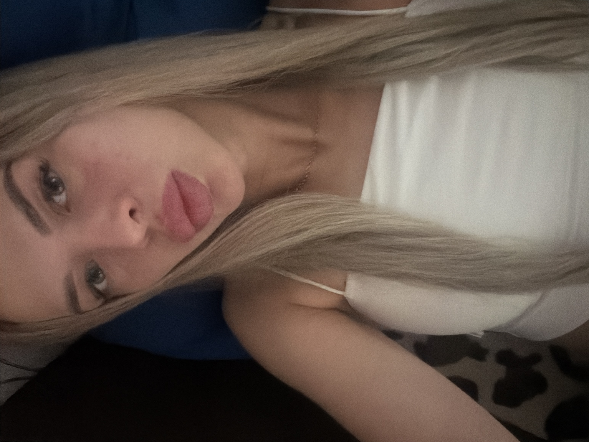 A close-up, selfie-style portrait shows a young woman with blonde hair. She is wearing a white tank top and a delicate gold chain necklace. Her facial features are prominent, with full lips and dark eyes. The background is dark and slightly out of focus, with what appears to be a blue cushion or pillow visible behind her. There are no discernible landmarks or contextual clues within the image to suggest a specific location in Kyiv, Ukraine, or any city. The image appears to be taken indoors in low lighting conditions.
