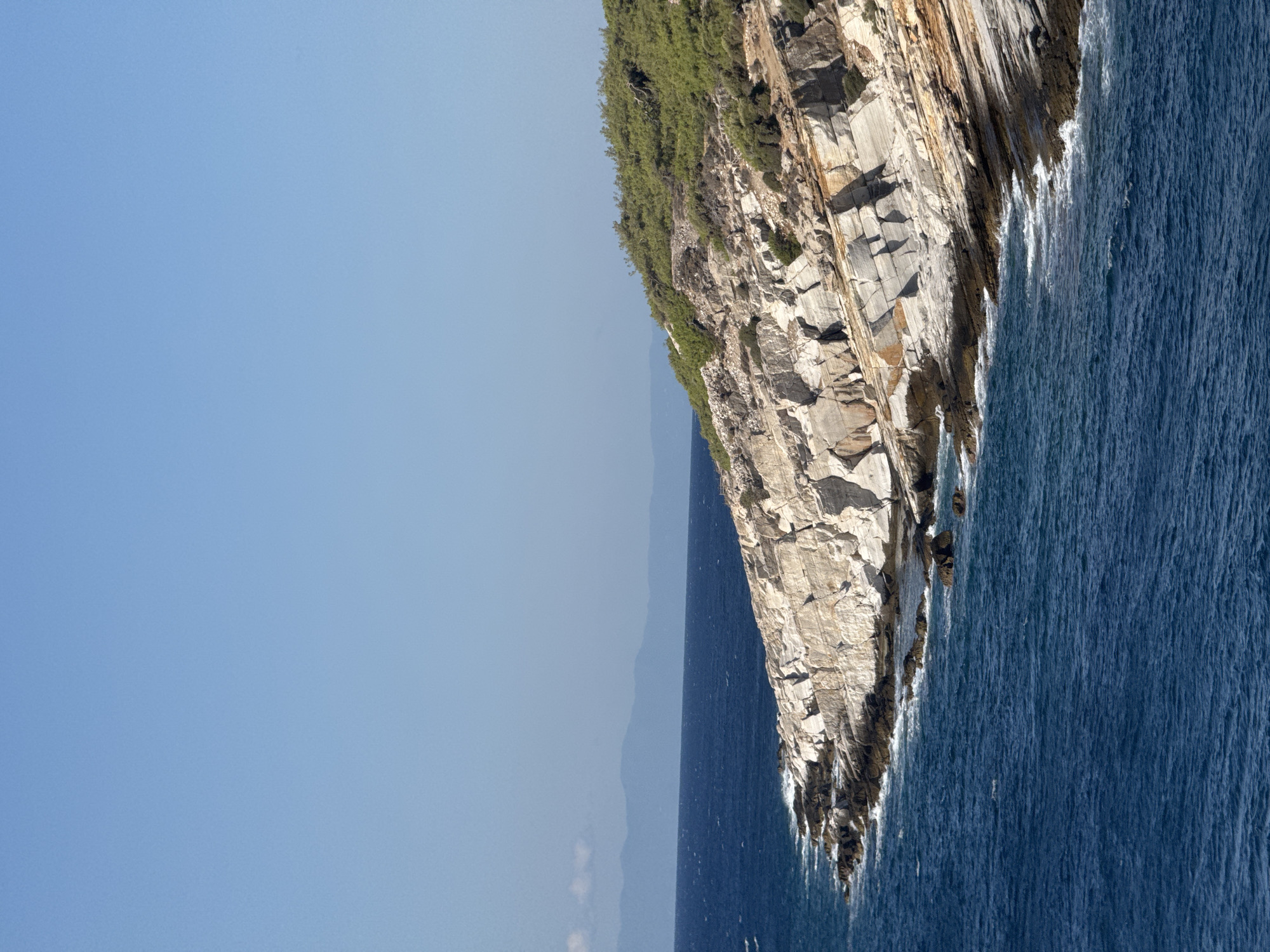 A vertical, eye-level, medium shot captures a coastal cliffside in Greece on a clear, sunny day. The deep blue sea, with gentle white waves lapping against the shore, dominates the lower half of the frame. The rocky cliff face, predominantly light gray and cream with streaks of orange and brown, rises sharply from the water, showcasing layered sedimentary rock formations. Green scrub and trees densely cover the top of the cliff, sloping upwards and to the right. In the far distance, a hazy mountain range is visible against the horizon. The sky is a clear, uniform light blue. There are no people or animals present in the image. The scene is serene and natural.