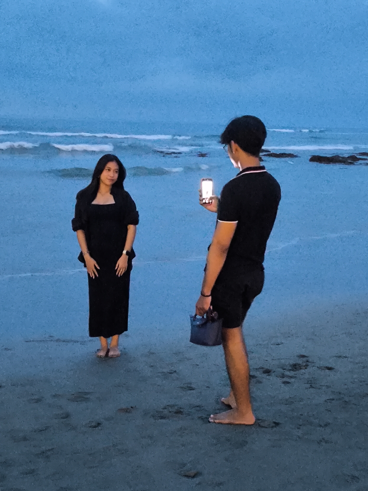 A young woman in a long black dress stands barefoot on a sandy beach, posing for a photograph. A young man, also barefoot, holds a smartphone up to take her picture. He is wearing a black polo shirt with white trim and black shorts, and he is holding a dark blue bag. The background shows the ocean at dusk, with visible waves and rocks in the distance. The location is the city of Canggu, Indonesia.