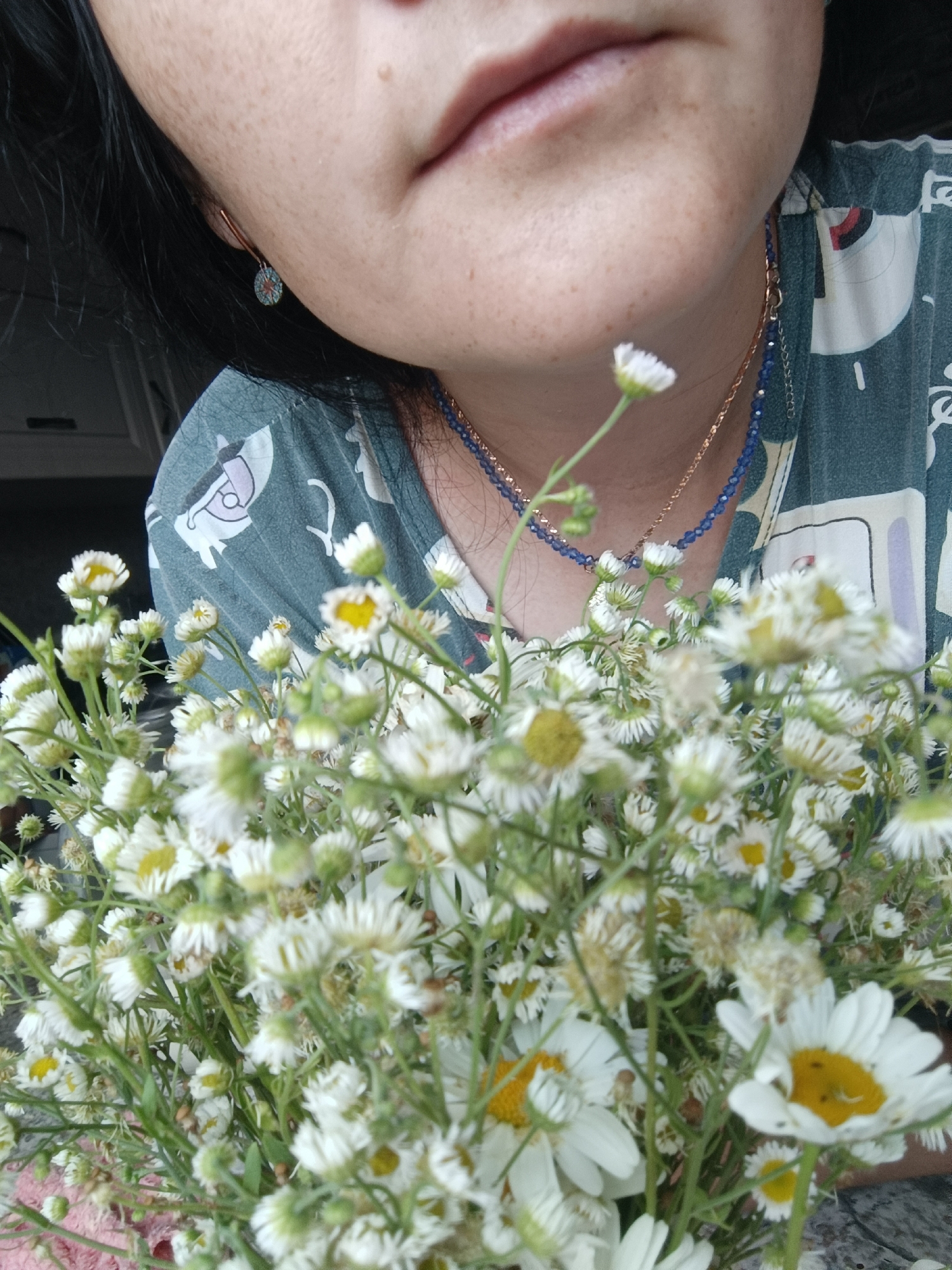 The image is a close-up shot of a person's face and a bouquet of small white flowers. The person's chin and mouth are visible, with a few freckles on their skin. They are wearing a dark green shirt with a white graphic pattern, a gold chain necklace, and a blue beaded necklace. An earring with a circular blue and red design is also visible. The flowers, likely daisies, are the primary focus of the lower half of the image, with many in bloom and some buds. The background is blurred, but appears to be indoors with a hint of a window or cabinet. There is no visible indication of the city of Karsha, Russia, or any specific environmental details that would place it there.