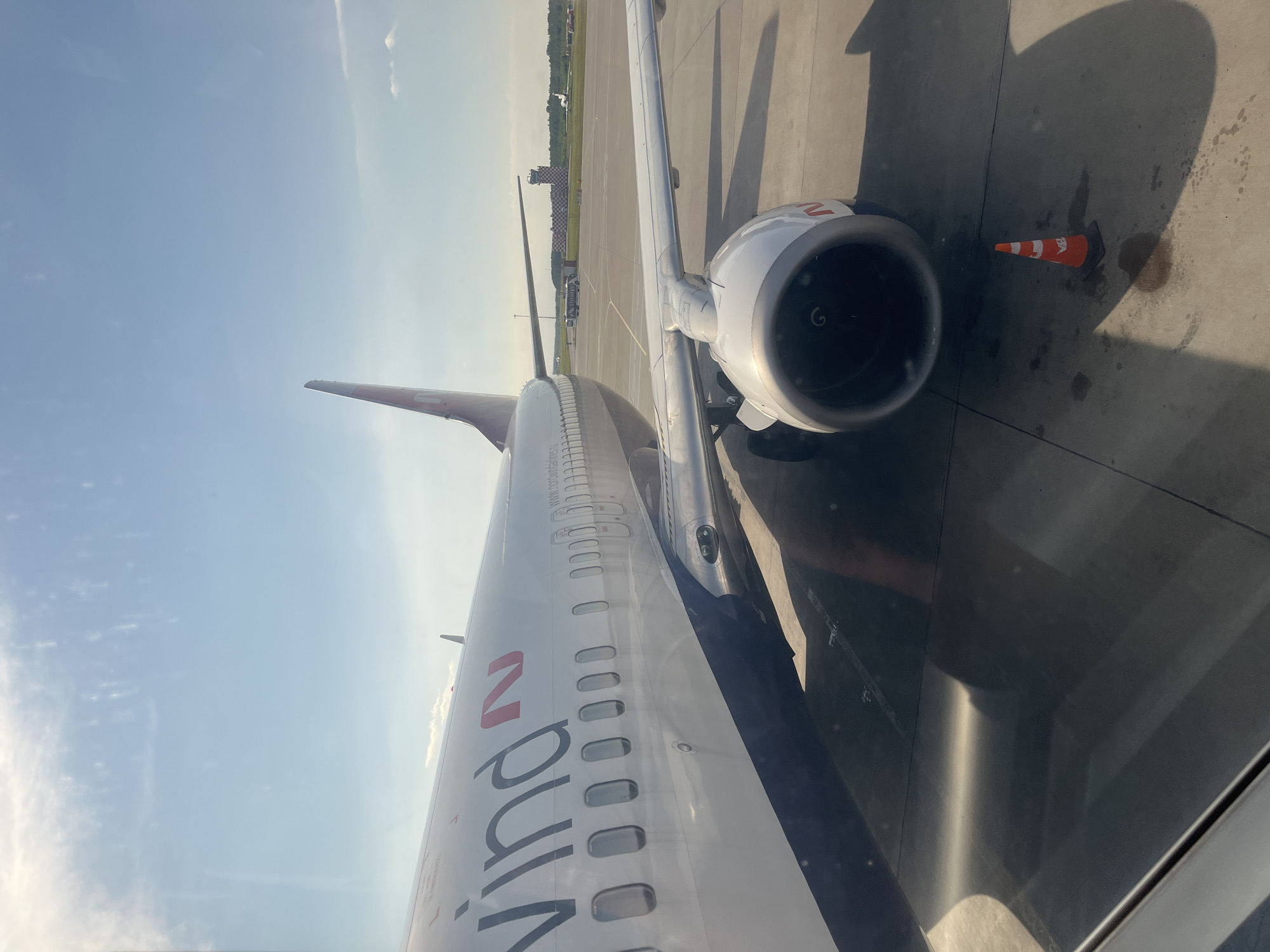An airplane is parked on the tarmac at Pulkovo Airport (LED) in Saint Petersburg, Russia. The plane, a Nordwind Airlines Boeing 737, is white with a dark blue stripe along the bottom of the fuselage and the airline's logo on the tail. The name "Nordwind" is visible on the side of the plane, along with the airline's website address. The image shows a portion of the wing and the engine, which is large and white with red accents. An orange traffic cone is placed on the tarmac near the engine. In the background, a control tower with a checkered pattern is visible, along with other aircraft and airport buildings. The sky is blue with some white clouds, indicating a sunny day. The image is taken from inside the airplane, looking out the window.