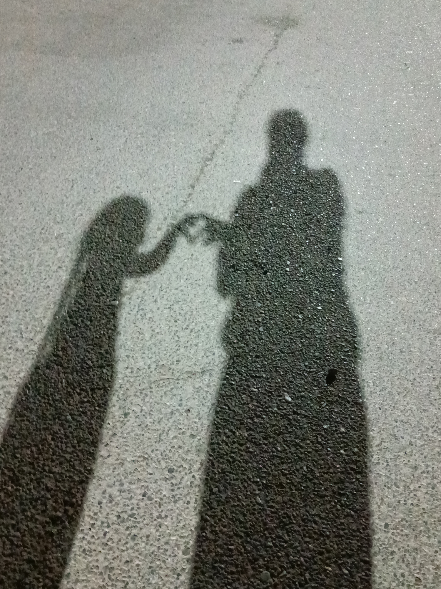The image shows the shadows of two people, one larger than the other, on an asphalt surface. The larger shadow, on the right, appears to be an adult, with a distinct head, torso, and outstretched arm. The smaller shadow, on the left, is tilted and appears to be a child or a person in a different pose, with their arm also extended. Their hands are clasped together, forming a heart shape. The asphalt is textured with small pebbles. The lighting suggests it is either dawn or dusk, casting long shadows. There are no discernible landmarks in the background or foreground to indicate a specific location within Shymkent, Kazakhstan.