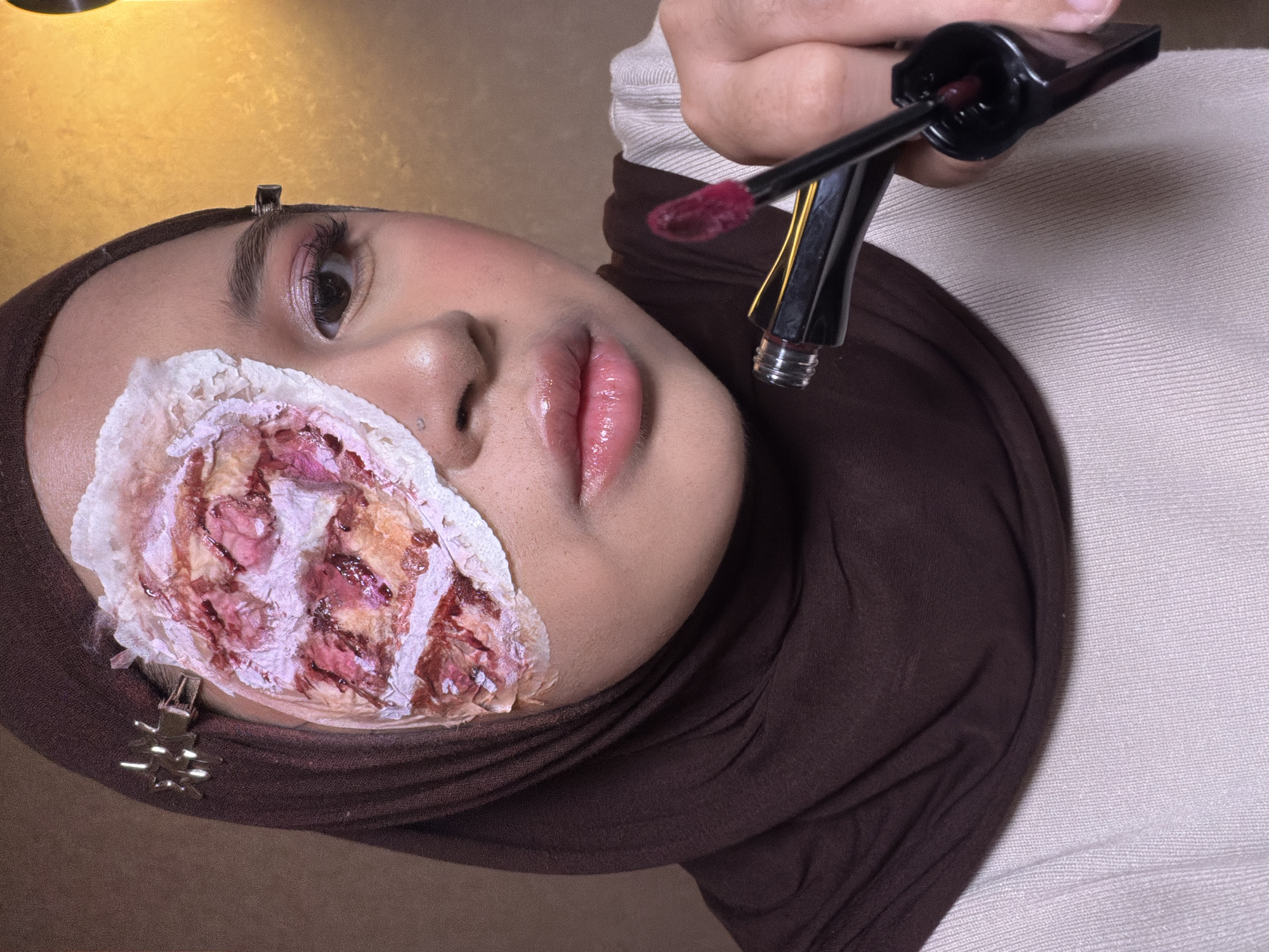 A person wearing a brown hijab is pictured applying lip gloss, while also having a gruesome special effects makeup applied to the left side of their face, resembling an open wound. Two hair clips are visible in the hijab, near the special effects makeup. The background appears to be a neutral-colored surface with soft, diffused lighting. The location of the image is not discernible as East Jakarta, Indonesia.