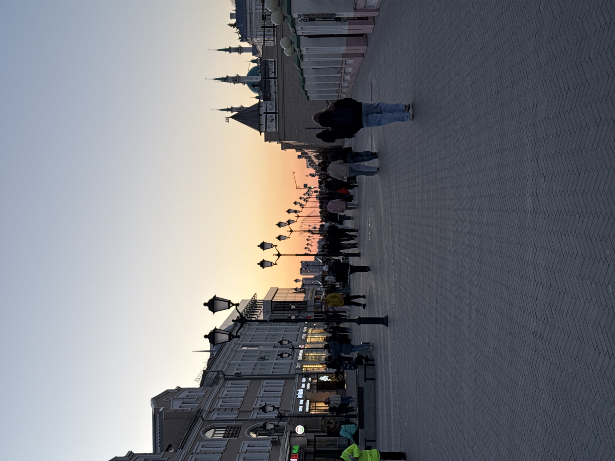 The image shows a view of Bauman Street in Kazan, Russia, at dusk. A pedestrian street lined with buildings on the left and the Kazan Kremlin in the distance to the right. A crowd of people walks along the street, which is paved with dark-colored bricks in a herringbone pattern. Street lamps are spaced along the street. The sky is transitioning from light blue to orange, indicating sunset.