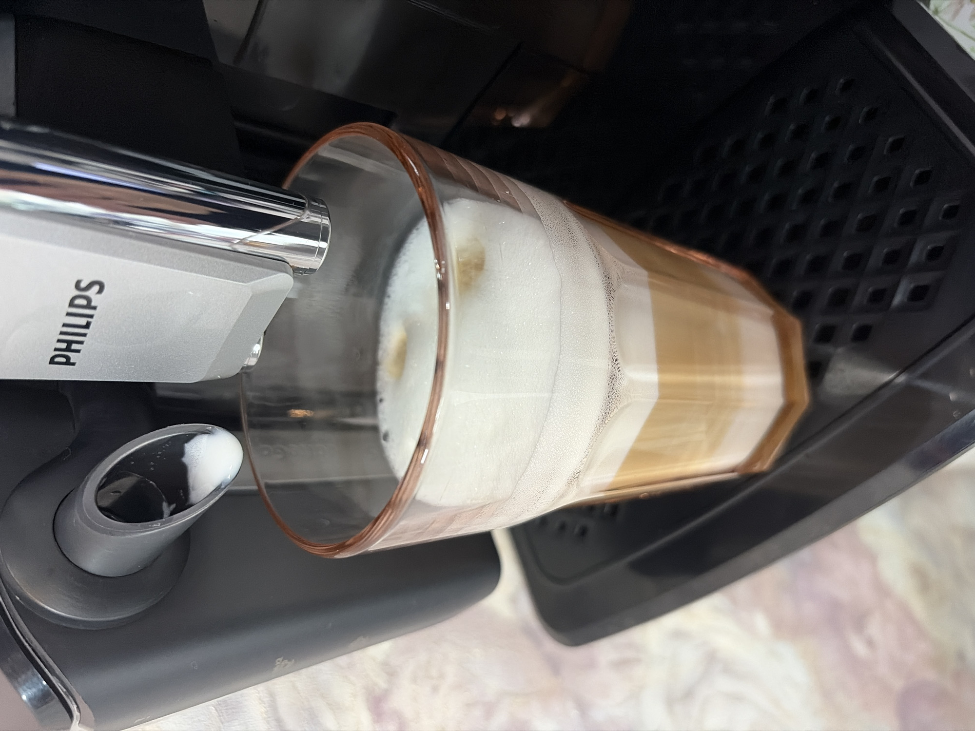 In Saint Petersburg, Russia, a close-up shot captures a Philips coffee machine dispensing a frothy beverage into a clear, faceted glass. The coffee, a layered mixture of dark brown and creamy beige, is topped with a thick layer of white foam. A small amount of white liquid, likely milk, is visible exiting the machine's spout, indicating the brewing process is underway or has just concluded. The coffee machine, primarily dark grey and metallic silver, is positioned over a black drip tray with a patterned surface. The background is softly blurred, hinting at a light-colored, possibly textured surface. The image composition focuses on the drink and the immediate interaction with the coffee machine.