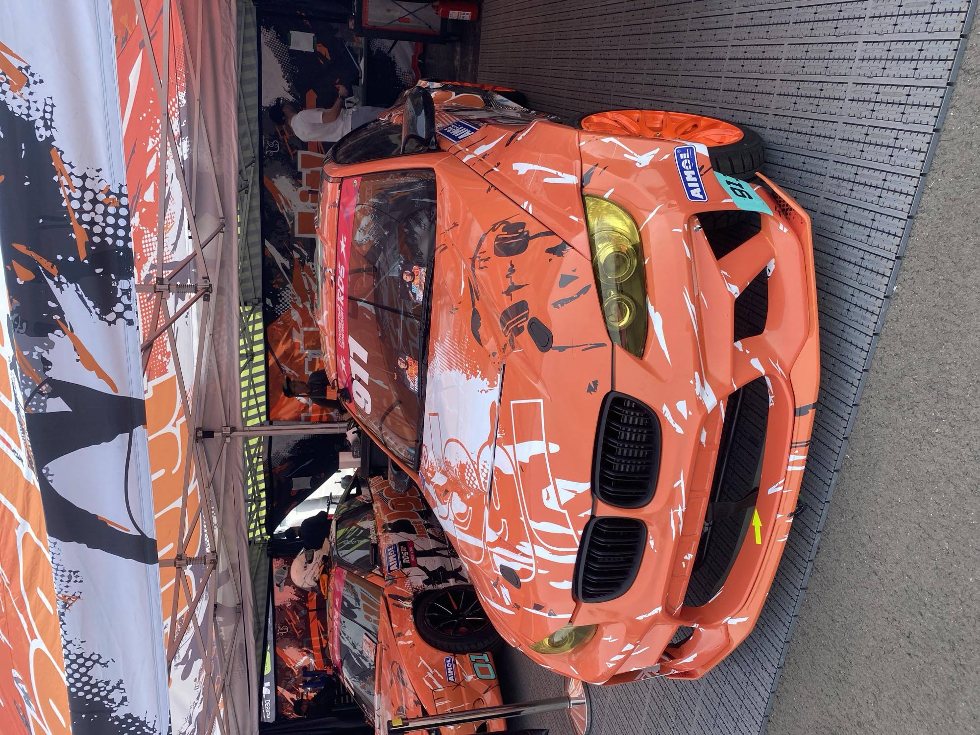 The image shows two custom-designed drift cars parked under a tent. The car in the foreground is an orange BMW with a white and black abstract graphic design. It has a wide body kit, yellow headlights, and orange rims. The car behind it is also orange and appears to be a Nissan, with a similar graphic design and the number "91" visible on its windshield. Both cars are heavily stickered with sponsor logos. In the background, people are seen standing around the cars, and there are tents and banners suggesting a racing or event setting. The ground appears to be a temporary flooring material laid over asphalt. The context provided places this scene in Minsk, Belarus.