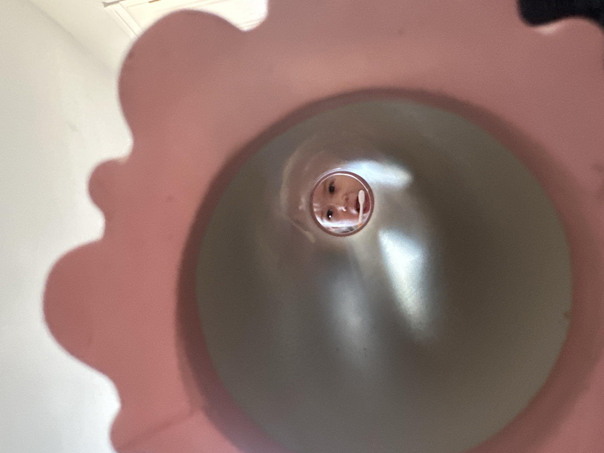 The image presents a close-up, upward-looking perspective into a large, pink, scalloped object, likely a toy or part of a play structure, which forms a tunnel or tube. At the far end of this tunnel, a child's face is visible, peeking into the opening. The child's eyes are looking directly towards the camera, and their mouth is slightly open, conveying an expression of curiosity or playfulness.
The inner surface of the tunnel is a smooth, light greyish-pink, reflecting light sources as bright streaks. The scalloped edges of the pink object fill a significant portion of the foreground and sides of the frame. Beyond the immediate pink object, a plain white wall and a white ceiling are visible, indicating an indoor setting. The lighting appears to be natural or artificial indoor light. There is no discernible text in the image. The scene captures a moment of childhood interaction with a play item in Kharnavy, Russia.