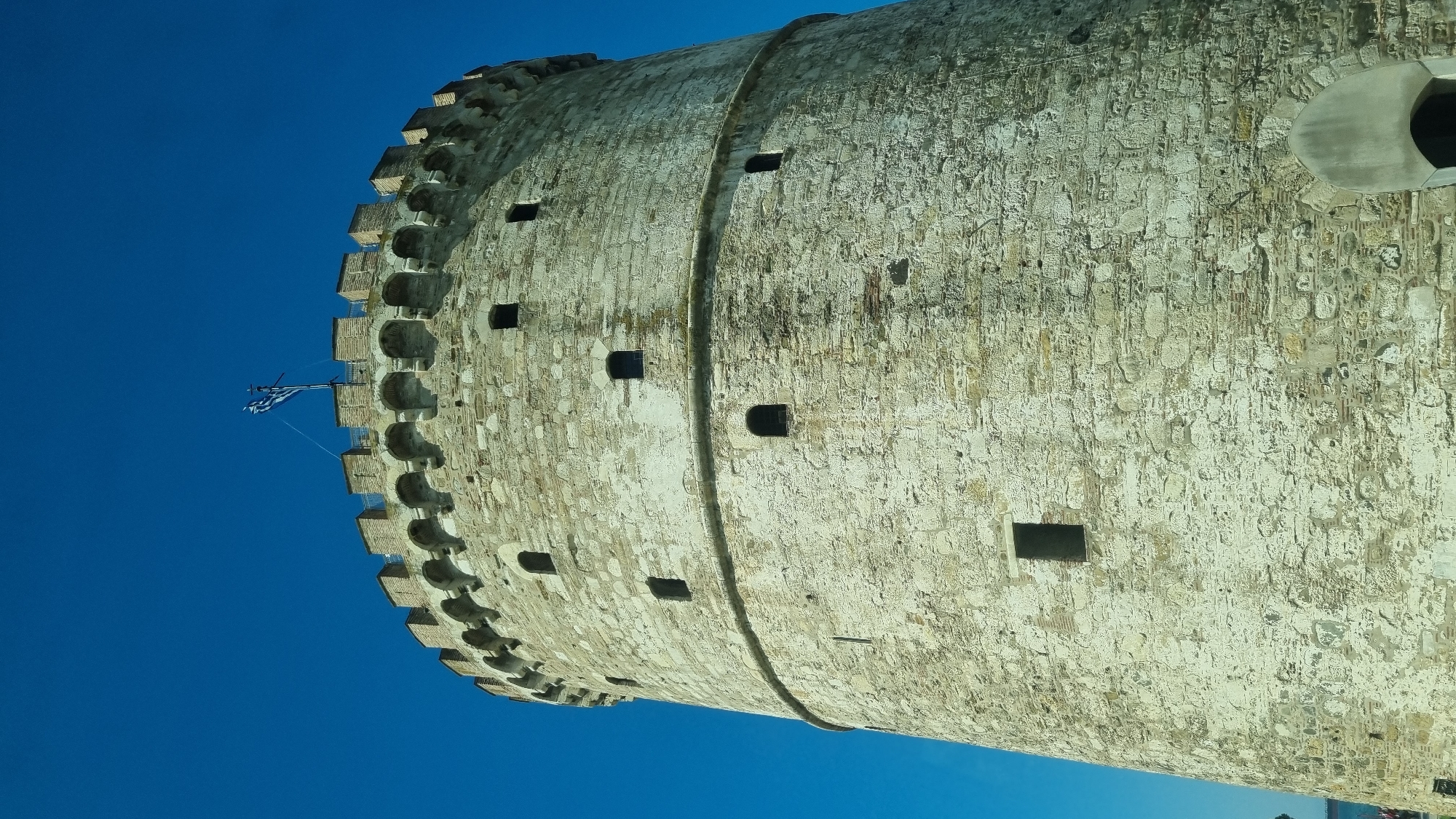 The image shows the White Tower, a historical monument and museum in Thessaloniki, Greece. The tower is a cylindrical stone structure with crenellated battlements at the top. A Greek flag flies from a flagpole on the battlements. Several small, dark windows are visible on the exterior of the tower. The tower is set against a clear blue sky. The bottom of the image shows a glimpse of a body of water and what appears to be a boat, suggesting the tower is located near the waterfront.
