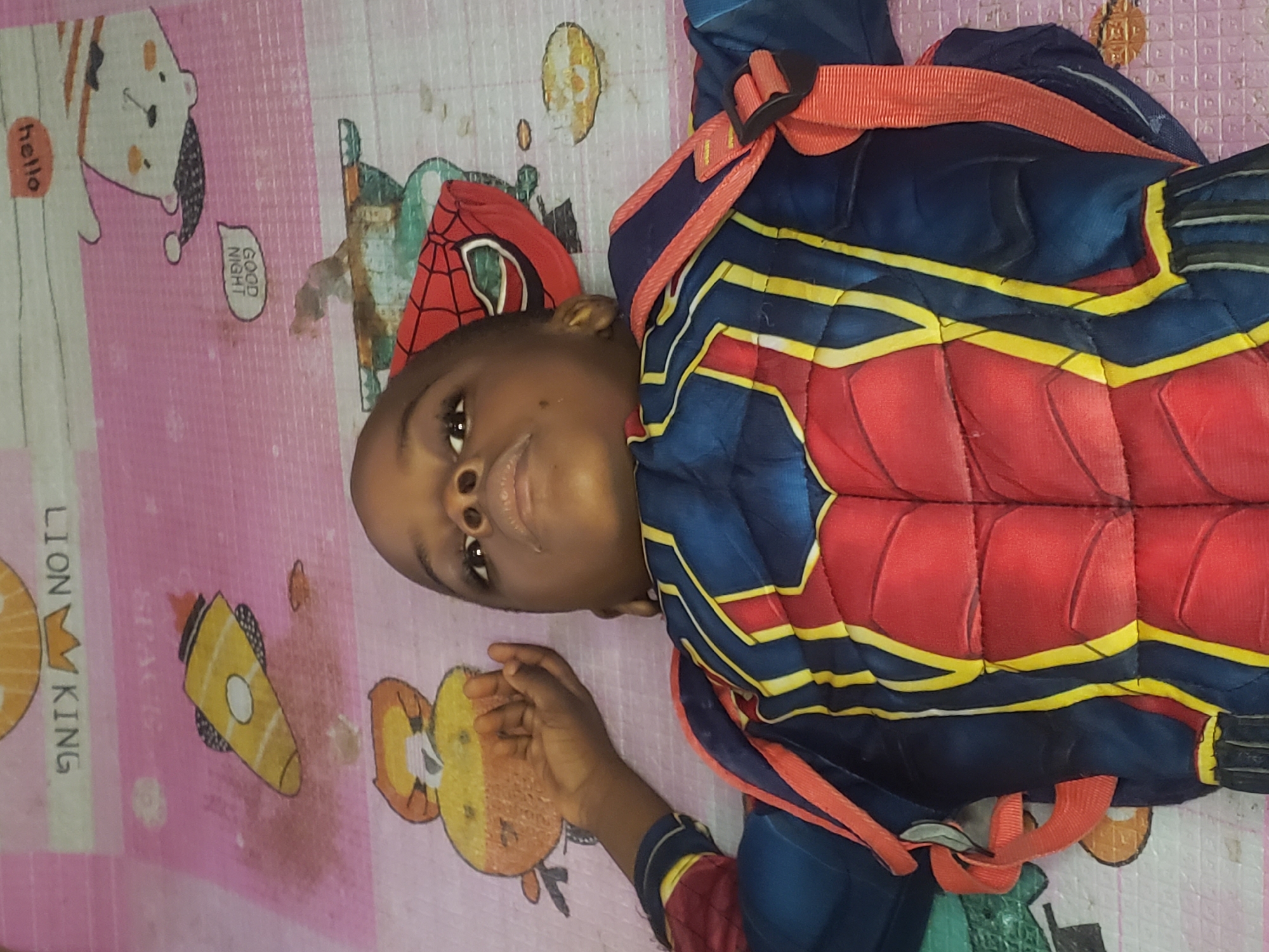 A young child, dressed in a blue and red superhero costume with yellow accents, lies on their back on a pink mat. The child's head is slightly tilted back, and their eyes are open and looking upwards. A red Spider-Man mask rests on the mat near their head. The mat has cartoonish illustrations of a rocket ship and a bear, as well as text that appears to read "HELLO" and "GOOD NIGHT." The image was taken in Narayi, Nigeria.