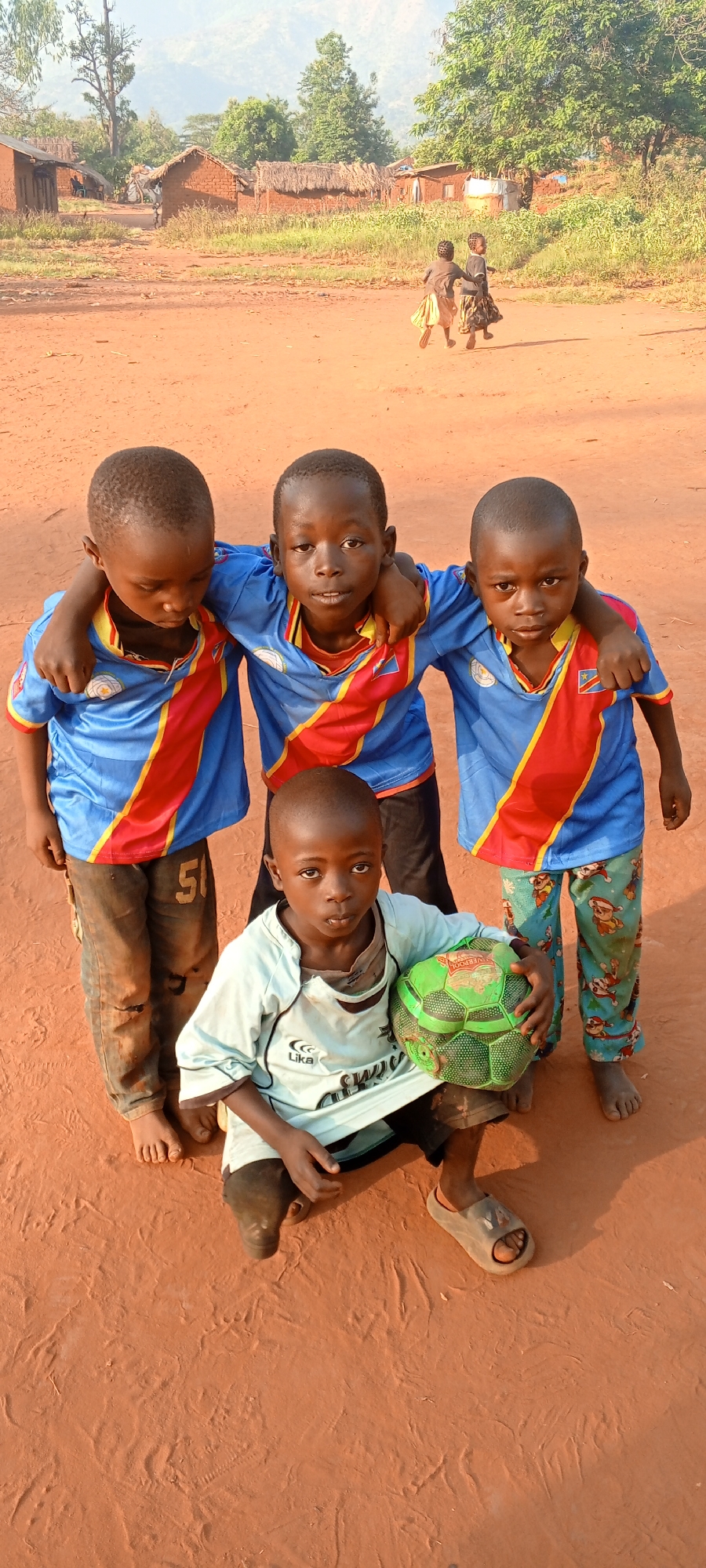 In Ngalula, Democratic Republic of the Congo, a group of four young children are posed in front of a rural dirt road. Three boys stand in the back row, arms interlocked across their shoulders. All three are wearing identical blue soccer jerseys with red and gold shoulder panels. The child on the left wears tattered blue jeans, and the boy on the right wears patterned pajama pants. A fourth child kneels in front, holding a green soccer ball, wearing a light blue soccer jersey. In the background, several brick and thatched-roof houses are visible. Two young children run in the far distance. The surrounding area is predominantly bare, red dirt.