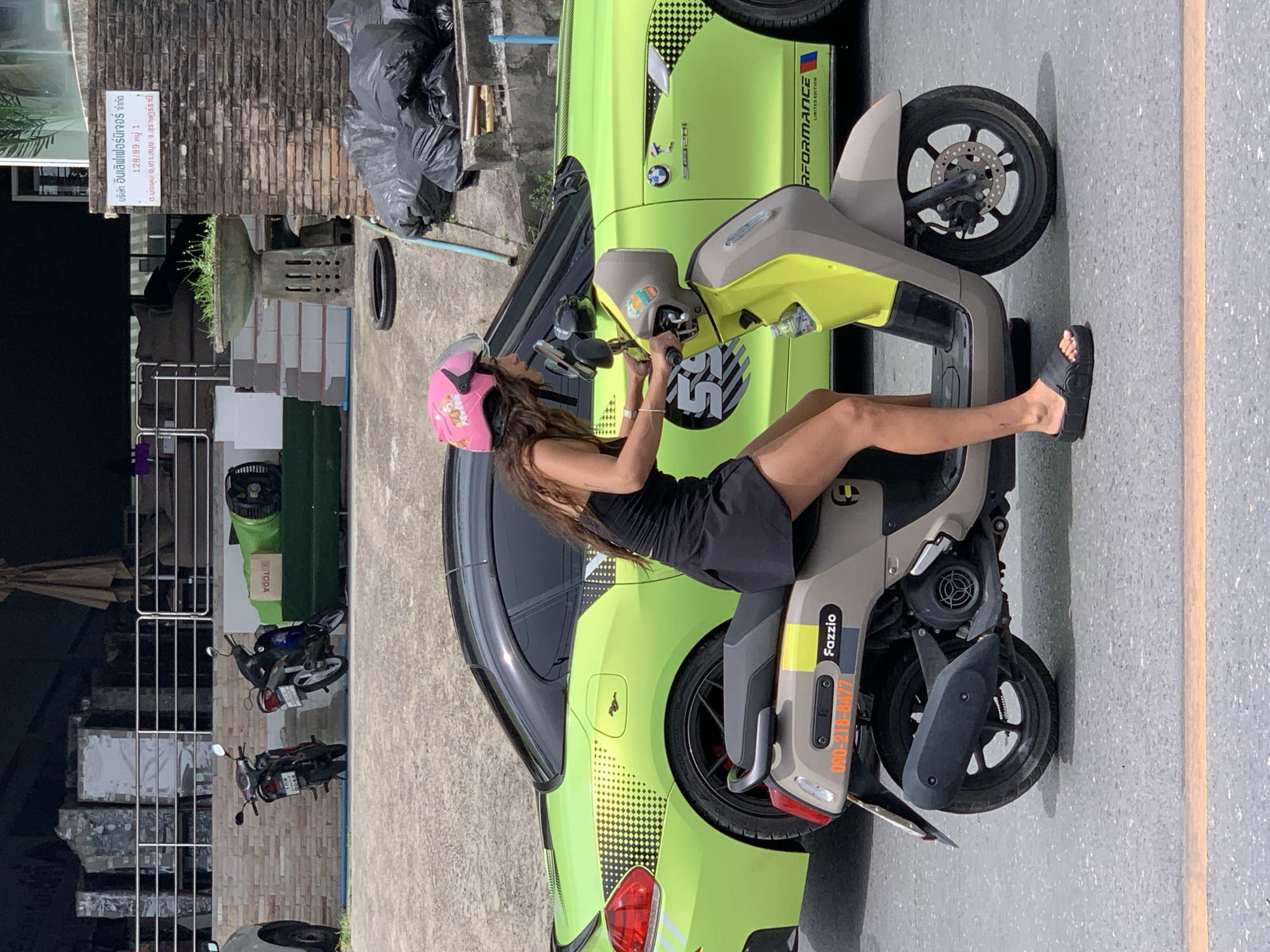 A woman wearing a pink helmet with a cartoon pig design and a black dress is sitting on a light brown and yellow scooter. She is holding the handlebars and looking forward. The scooter has a black seat and black wheels. Behind the scooter is a bright green car with black accents and a black roof. The car has a graphic design on the side that includes a checkered pattern and a horse silhouette. In the background, there are two motorcycles parked, a building with brick walls, and stacks of what appear to be large, rectangular objects covered in plastic. There are also black trash bags piled up and a concrete planter with greenery. The scene appears to be on a paved road with a yellow line. The location is identified as Baan Bo Phut, Thailand.