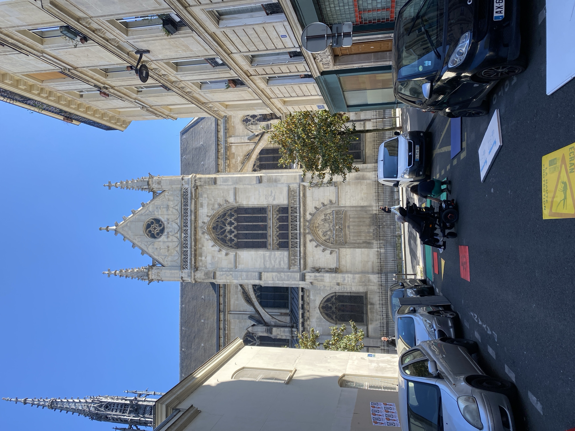 This image captures a bright, sunny daytime scene on a city street in Paris, France, with a prominent gothic-style church dominating the background.
The focal point is the large church, likely made of light grey stone, featuring intricate architectural details such as pointed spires, rose windows with dark tracery, arched entrances, and decorative carvings. Its tall structure is set against a clear, deep blue sky. Another tall, slender spire is visible further in the background to the left.
In the foreground and midground, a narrow street is lined with parked vehicles and classic Parisian buildings. On the right side of the street, several cars are parked bumper-to-bumper, including a black Smart car with a French license plate "AK-6..." and a white van displaying "ES-711-OG". Further along, a silver hatchback is also visible. Rectangular patches of color (red, green, blue) are painted on the asphalt next to the parked cars, possibly indicating specific zones.
A person is seated in a black motorized wheelchair on the street, near the parked vehicles, being attended to by another individual. A small tree with green leaves stands between the street and the church facade.
The buildings flanking the street exhibit typical Parisian architecture with light-colored stone facades, numerous windows, and decorative cornices. A shopfront with a dark green awning and a circular street sign are visible on the right.
Notable details include a yellow warning sign on the pavement to the right, featuring a mosquito icon and the French text "ÉCRAN HAUTE PROTECTION" (high protection screen) and "MOUSTIQUE" (mosquito). Partial street markings, possibly indicating "PAYANT" (paid parking), are also visible. The bright conditions suggest it is midday or early afternoon.