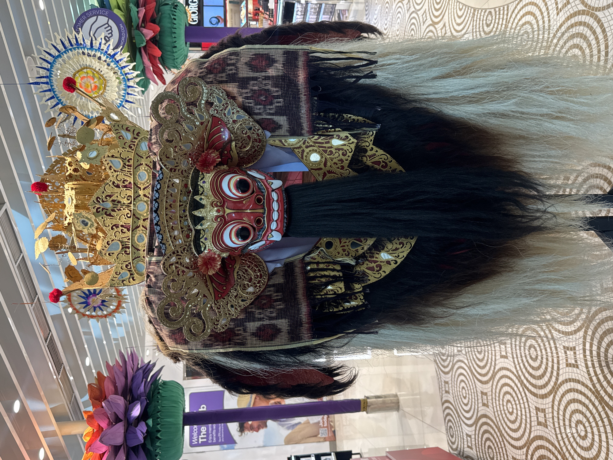 A vibrant, intricately designed Balinese Barong mask is prominently displayed, its red face with large white eyes and black pupils gazing forward. Sharp fangs protrude from its mouth, and a dark mustache-like feature is visible. The mask is adorned with an ornate golden headdress featuring delicate patterns, mirrors, and red pom-poms. Dark and light shaggy hair or fur cascades from the sides and top of the mask. Behind the mask's head, there's a patterned fabric with geometric designs.
The mask is part of a larger decorative display within what appears to be a modern indoor commercial space, possibly an airport or shopping center in Kuta, Indonesia. In the background, hanging from a high ceiling with white slatted panels, are several decorative elements. To the upper left of the mask, a large, circular white and blue decoration with a yellow-orange center is visible, reminiscent of a mandala or traditional sunburst. Below it, a vibrant, multi-colored lotus-like structure in shades of orange, purple, and green adds to the festive atmosphere.
The floor features a distinctive geometric pattern of interlocking light brown circles. Visible text on signs in the background includes "CUSTOMER SERVICE" and a partial website address ending in ".com". Another sign shows "GIORG" (likely part of a brand name), and a purple stand has text like "Welcome The b" and "Enjoyment benefits wo" along with "Join today" in a speech bubble. The scene is brightly lit by artificial lighting.