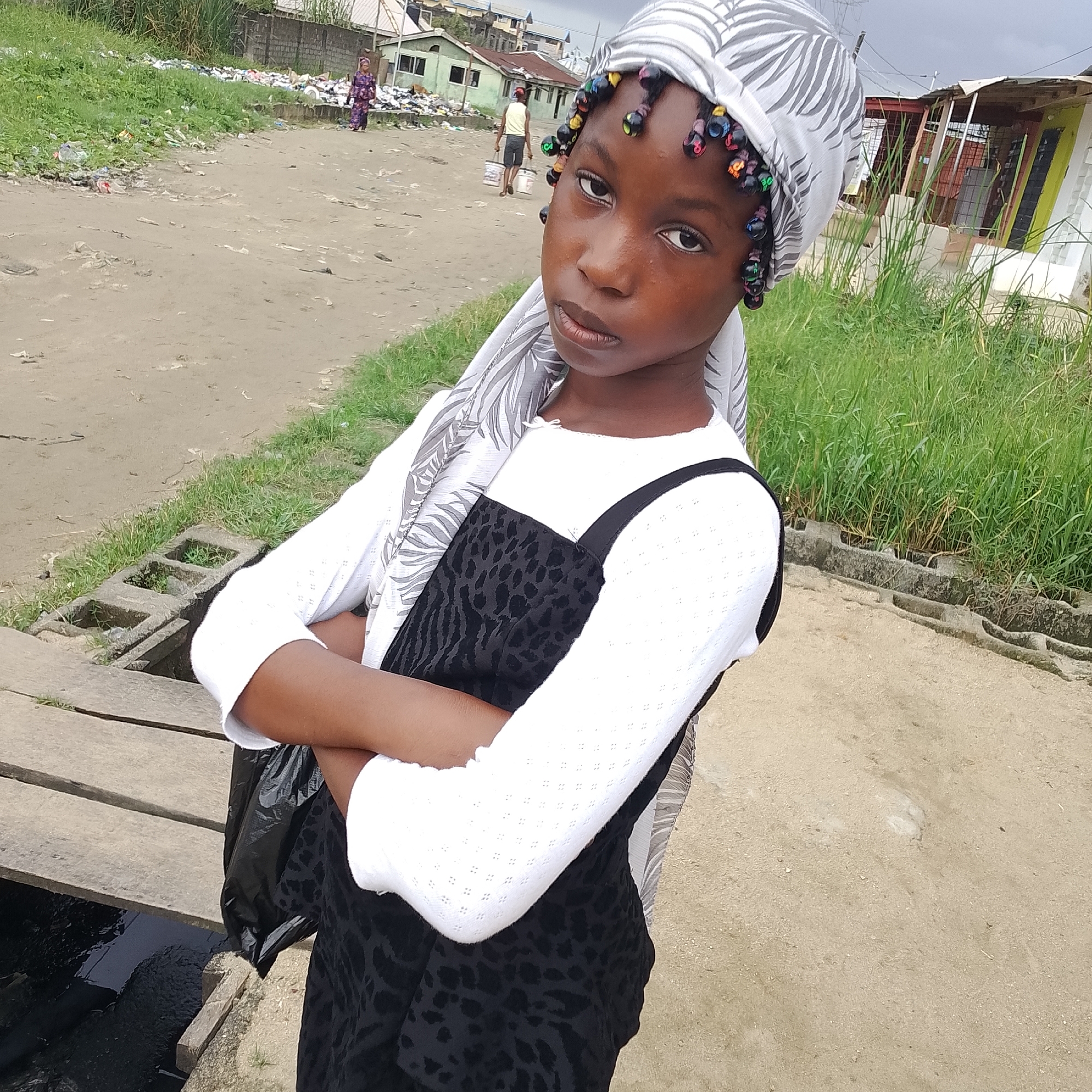 A young Black girl wearing a white and grey patterned headwrap and a white long-sleeved shirt over a black dress with a leopard print pattern stands with her arms crossed, facing the camera. Her hair is styled in braids adorned with colorful beads. The background depicts a dusty, unpaved street in Ado, Nigeria, with sparse vegetation and several small, simple buildings. Trash is visible scattered along the side of the street and in the background, indicating a potentially unhygienic environment. Two other individuals are visible in the distance: a woman in a patterned dress on the left and a man carrying two white buckets in the center. A wooden plank extends over a dark, murky drainage ditch in the foreground. The sky is overcast.