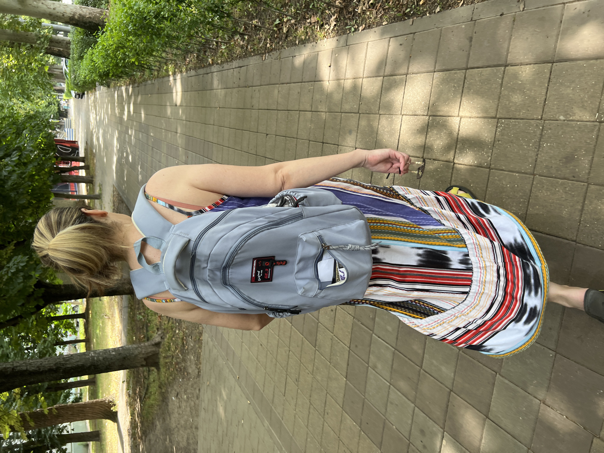 A woman with blonde hair pulled into a ponytail is walking away from the camera on a paved pathway in a park or tree-lined street. She is wearing a light blue backpack and a long, colorful dress with vertical stripes. In her right hand, she is holding a pair of sunglasses. The pathway is made of square concrete tiles and is flanked by lush green bushes on the right and trees on the left. The sun casts dappled shadows on the path. In the background, there are more trees, a glimpse of buildings, and parked cars. This scene is consistent with a typical urban park or boulevard in Krasnodar, Russia, particularly during warmer months.