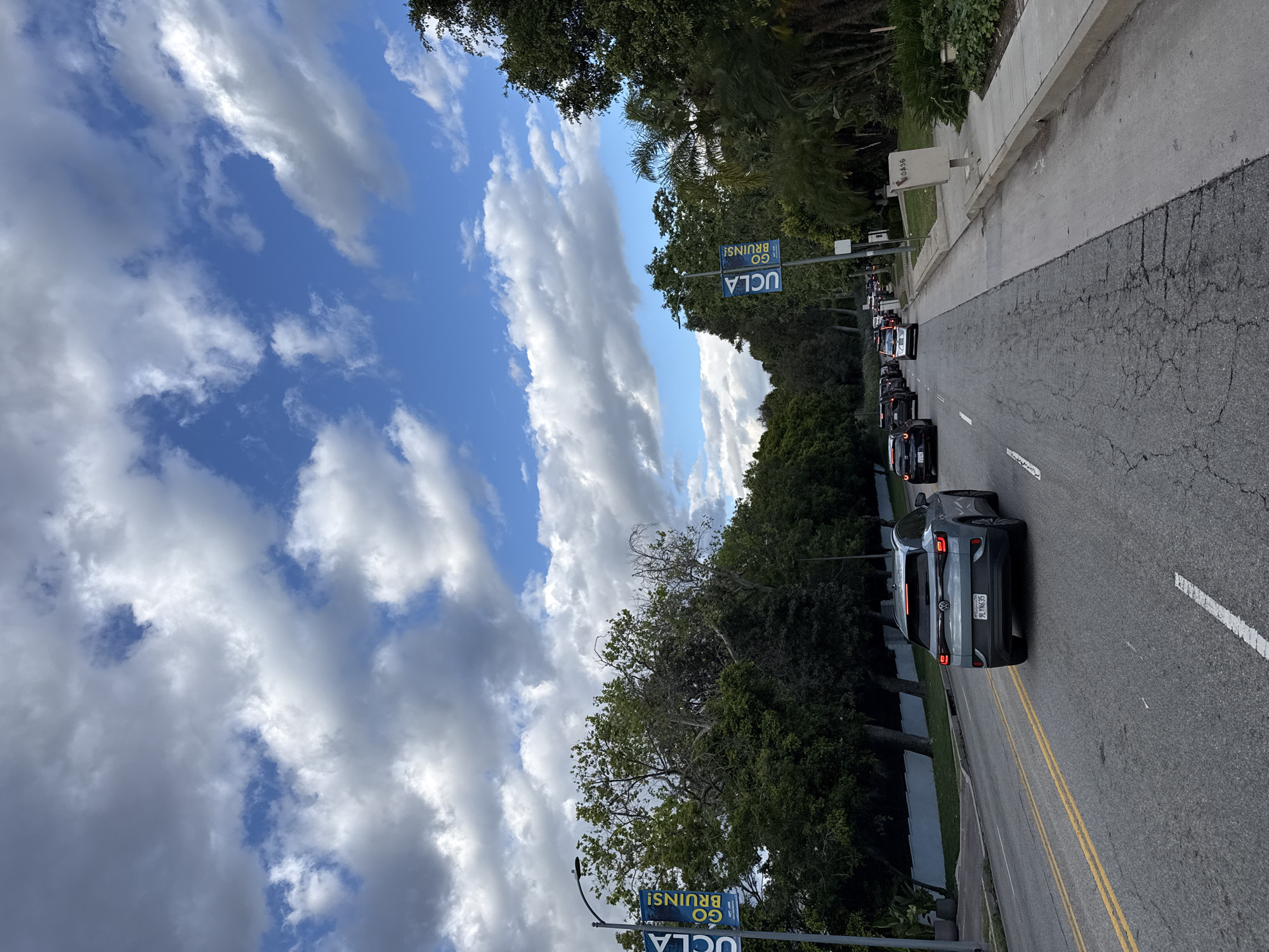 The image depicts a street in Los Angeles, California, with a line of cars moving slowly. The sky is visible with a mixture of blue sky and white clouds. "UCLA GO BRUINS!" banners are posted along the roadside. The road is asphalt, and there is a sidewalk on the right side of the road. A license plate is visible in the bottom right corner. There are trees and greenery along the side of the road.