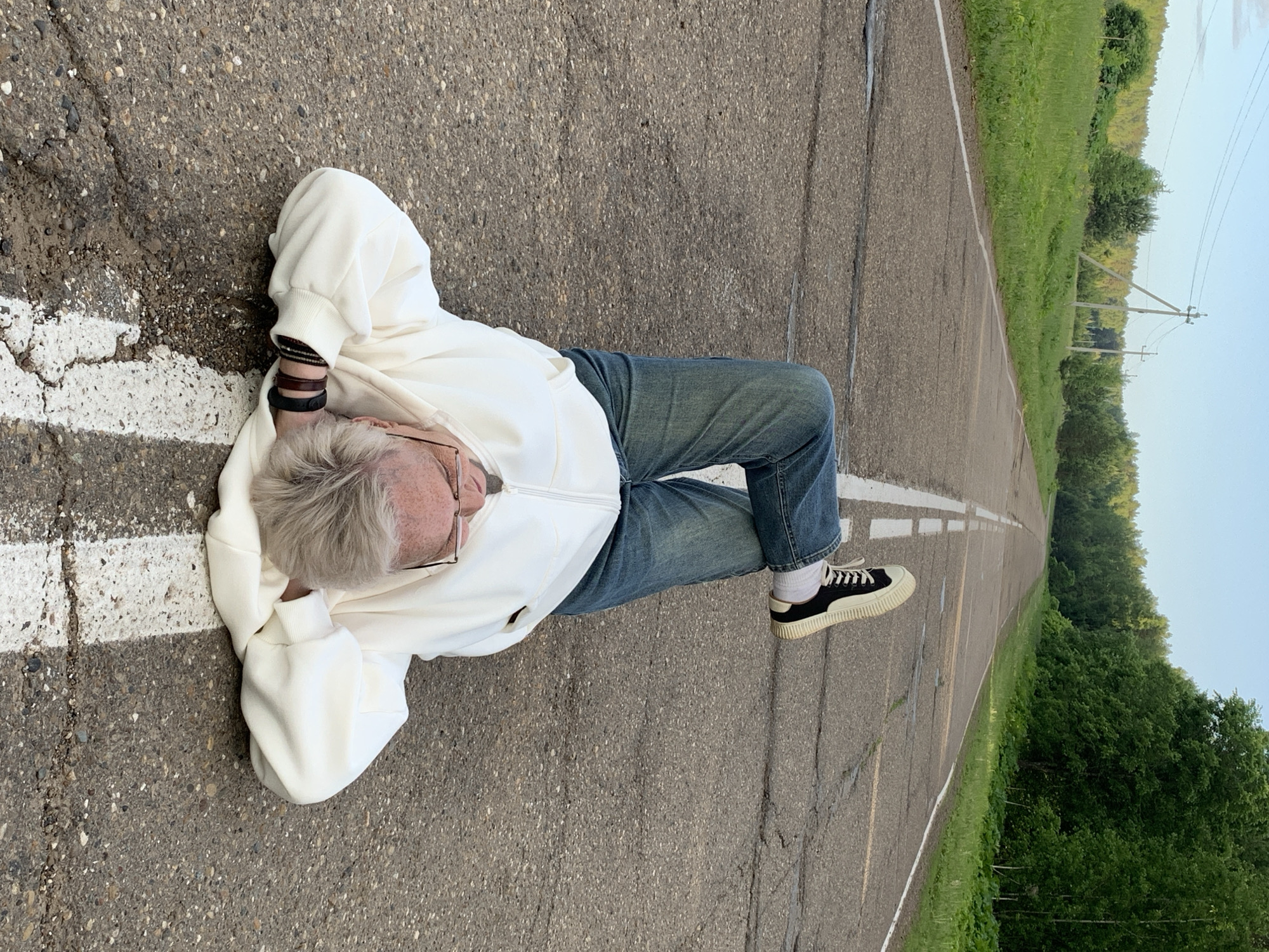 The image shows a person lying on their side on an asphalt road. The photograph itself is rotated 90 degrees clockwise from a natural horizontal perspective. The subject is an individual with short, light grey or blonde hair, wearing glasses, a white long-sleeved zipped jacket or hoodie, blue jeans, and black and white platform sneakers with white socks. Several dark bracelets are visible on their left wrist. Their head is resting on their arm, and they appear to be in a relaxed pose.
The asphalt road beneath them is grey, exhibiting some cracks and wear. A solid white line runs along the edge of the road in the foreground, with a broken white line visible further in the distance. To the right of the road, a lush green landscape of grass and trees extends into the background. Utility poles with visible wires are spaced along the edge of the greenery, receding into the distance. The sky, visible in the upper right quadrant of the image, is a clear, light blue, indicating daytime with soft, diffused lighting. No discernible text is present in the image. The setting suggests a rural or semi-rural location, consistent with Bol'shoi Bukor, Russia.