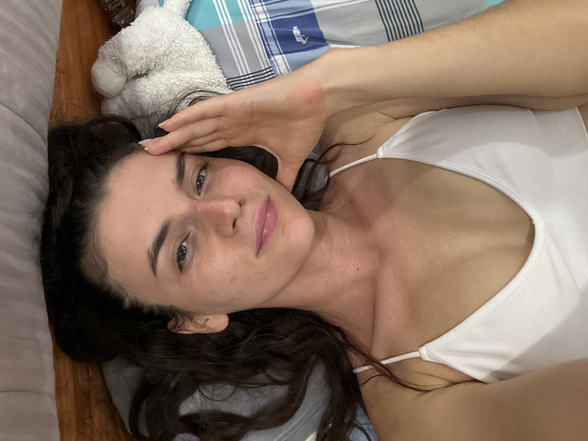 A woman lies on a bed with blue and gray plaid sheets in a brightly lit indoor space. She wears a white tank top. Her dark, wavy hair is spread on a gray pillow. Her right hand touches her forehead. To the left of her head is a white stuffed animal. It is unknown if the location is in Mũi Né, Vietnam.