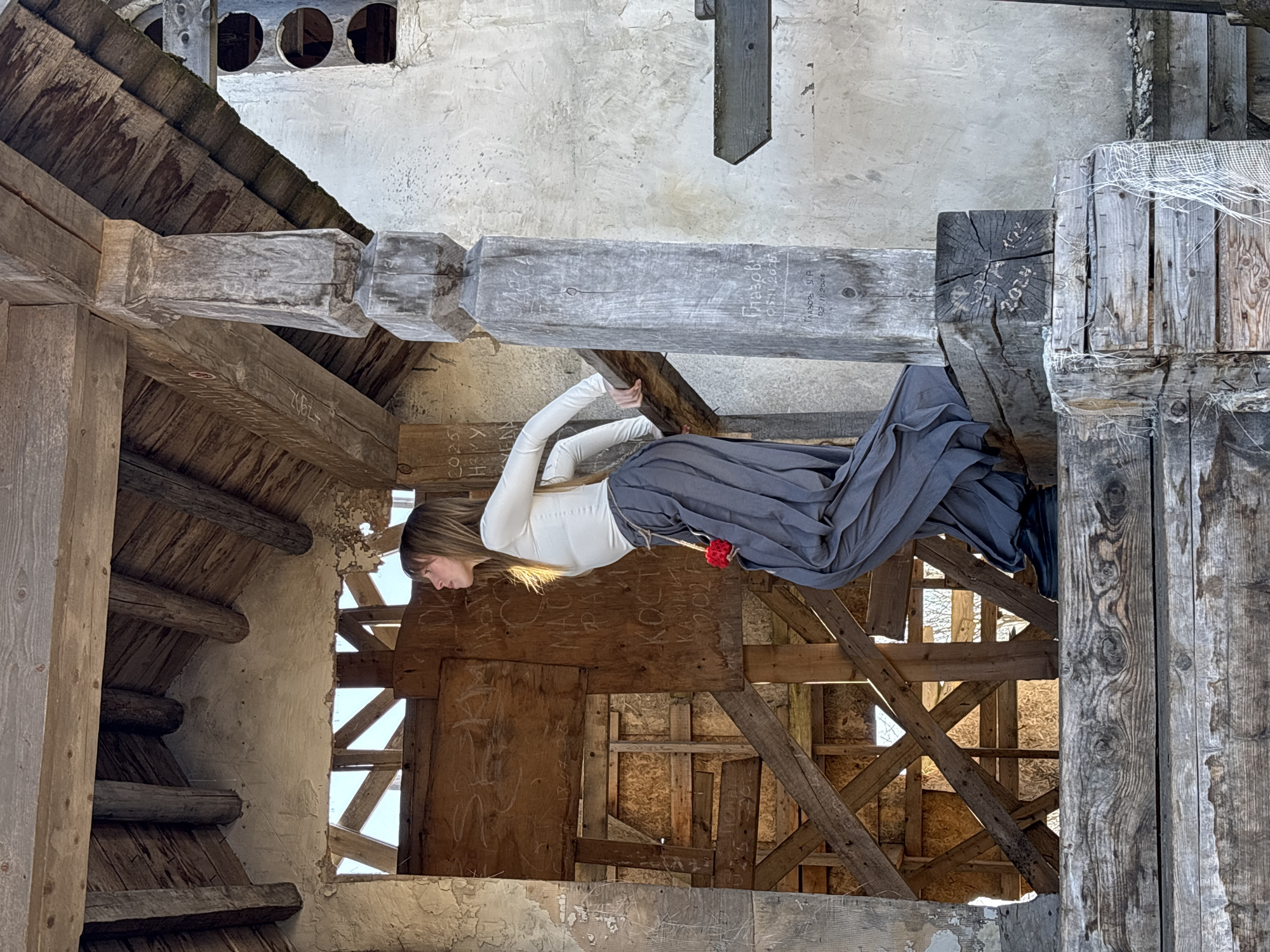 A woman with long, light brown hair stands inside a dilapidated wooden structure. She wears a white long-sleeved top and a long, grey skirt, with a red accessory hanging from her waist. The structure, heavily weathered with exposed beams and crumbling walls, appears to be an old building. The presence of graffiti on some of the wooden surfaces suggests possible human interaction or previous occupancy. The setting is the town of Gubakha, Russia, as indicated by graffiti on wooden structural elements.