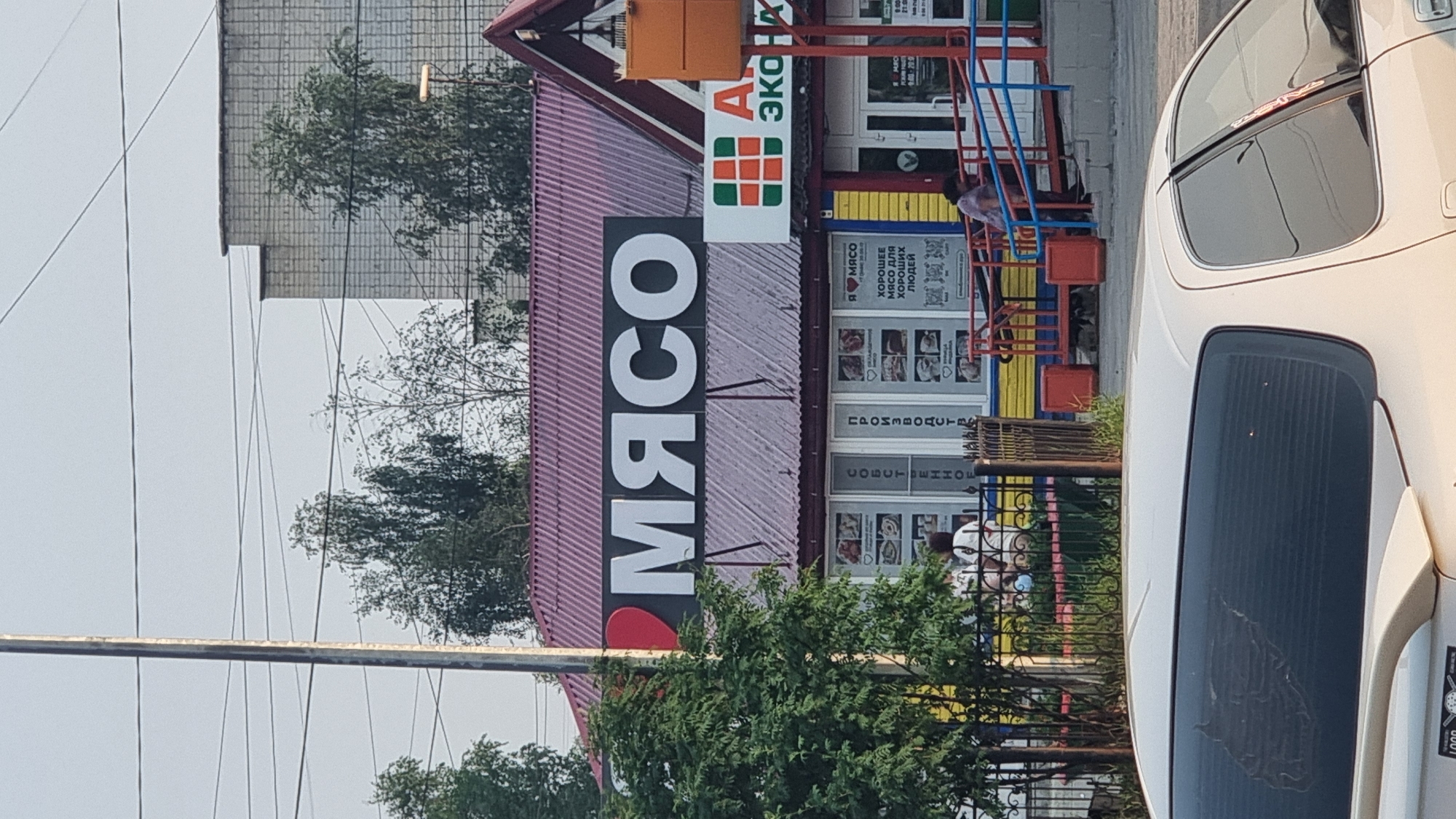 This is a medium-angle, exterior shot of a building with a large sign that reads "МЯСО" (Meat) in white lettering with a red heart icon. The building appears to be a store or market, indicated by storefront windows displaying images of food and text. Above the main sign, another sign features the letters "A ЭКОН ЭКА" which might suggest an ecological or economic focus.
In the foreground, a white car is partially visible, parked to the right of the building. The scene is outdoors, with trees and greenery present. A set of metal stairs, painted blue and red, leads up to the entrance of the building, and a person is seen sitting on the stairs. Another person is visible inside one of the windows. The lighting suggests it is daytime, with overcast skies. The overall impression is that of a local food establishment in a town or city setting. The text on the storefront windows includes phrases like "Я мясо" (I love meat), "ХОРОШЕЕ МЯСО ДЛЯ ХОРОШИХ ЛЮДЕЙ" (Good meat for good people), and what appear to be operating hours.