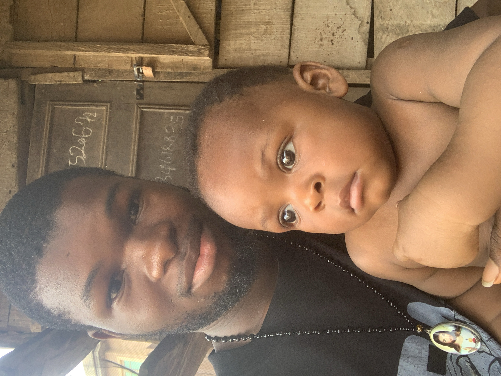 A close-up, portrait-oriented image features a young adult male and a baby, possibly captured as a selfie. The adult male, with dark skin, short dark hair, and a short beard, wears a black top and a black beaded necklace with an oval pendant depicting a religious figure, likely Jesus Christ, with a halo and the Sacred Heart. He looks towards the camera with a neutral expression. The baby, also with dark skin and short dark hair, is positioned next to him, seemingly leaning on his shoulder or chest, appearing bare-chested. The baby has wide, observant eyes and a slightly open mouth, also looking towards the camera.
The background reveals a rustic, wooden structure, consisting of rough wooden planks and a dark wooden door, consistent with a setting in Nkpor, Nigeria. Bright natural light suggests the photo was taken during the daytime. On the wooden door, there are two sets of numbers written in light chalk or paint: "52642" on the left and "34618835" on the right. The scene captures a moment of connection between the two subjects.