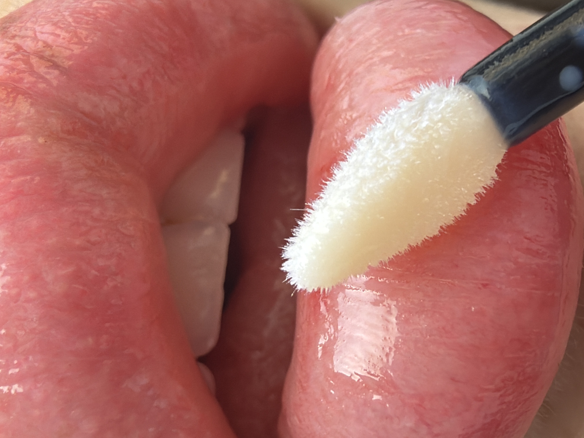 A close-up shot depicts a person applying lip gloss with a foam applicator to their lips. Two upper teeth are visible behind the lower lip. The lip surface shows a glossy sheen. The applicator has a black handle and a white sponge tip. The background is out of focus. There are no elements to indicate the location of Saint Petersburg, Russia.