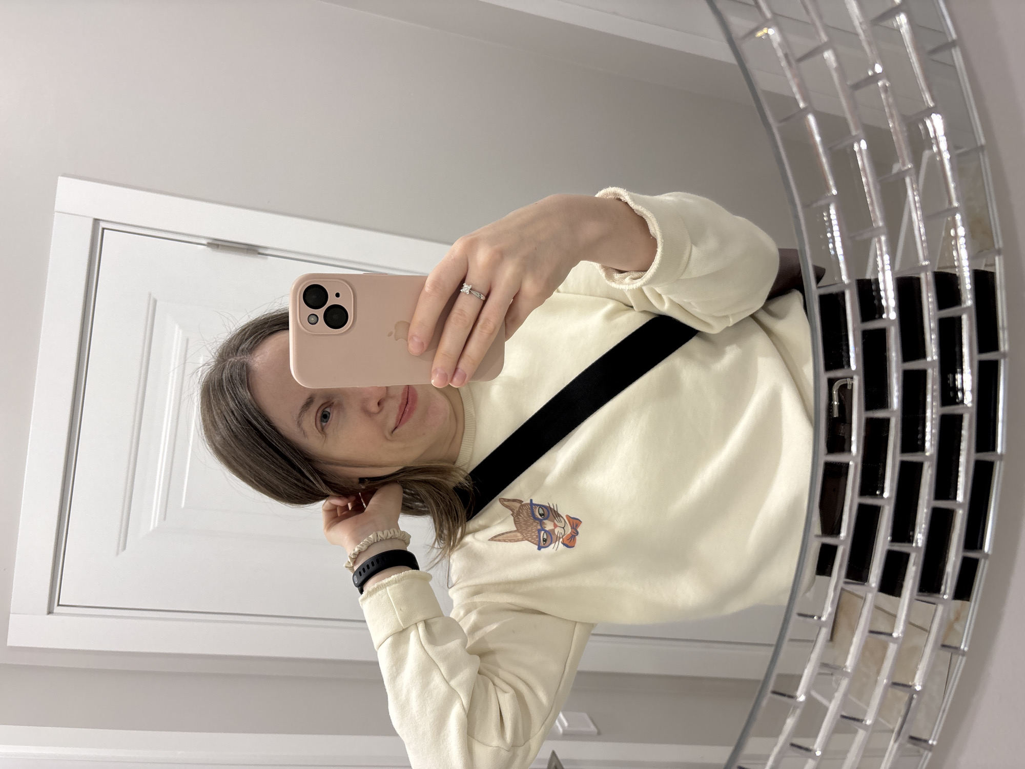 A woman stands in front of a mirror, using her smartphone to take a selfie. She has shoulder-length brown hair and is wearing a cream-colored sweatshirt with a black strap across her chest. The sweatshirt features a graphic of a cat wearing glasses and a bow tie. The woman has a ring on her finger and is wearing a black smartwatch on her wrist. The mirror is round and has a mosaic design around the edge. Behind her, there is a white door. The location is likely indoors, possibly a bathroom. The image does not provide information to verify it was taken in Saint Petersburg, Russia.