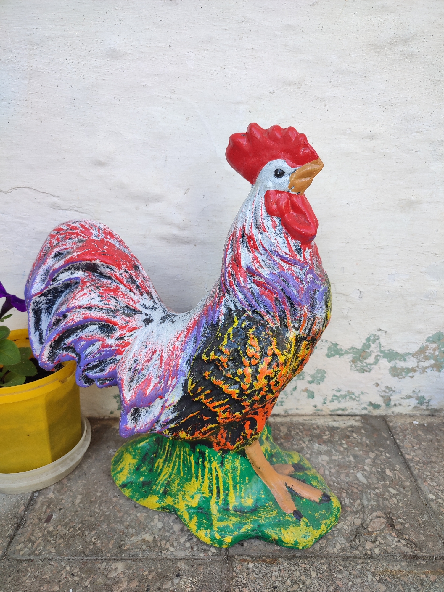 A colorful, three-dimensional rooster statue stands on a green and yellow base, positioned on a textured, light-colored tiled floor. The statue is detailed with various colors, including red, purple, black, orange, and white, with a red comb and wattle. To the left of the rooster, a bright yellow flower pot containing a plant with purple flowers is visible. The background is a rough, white wall with areas of peeling paint. This scene is located in the city of Voskobiinyky, Ukraine.
